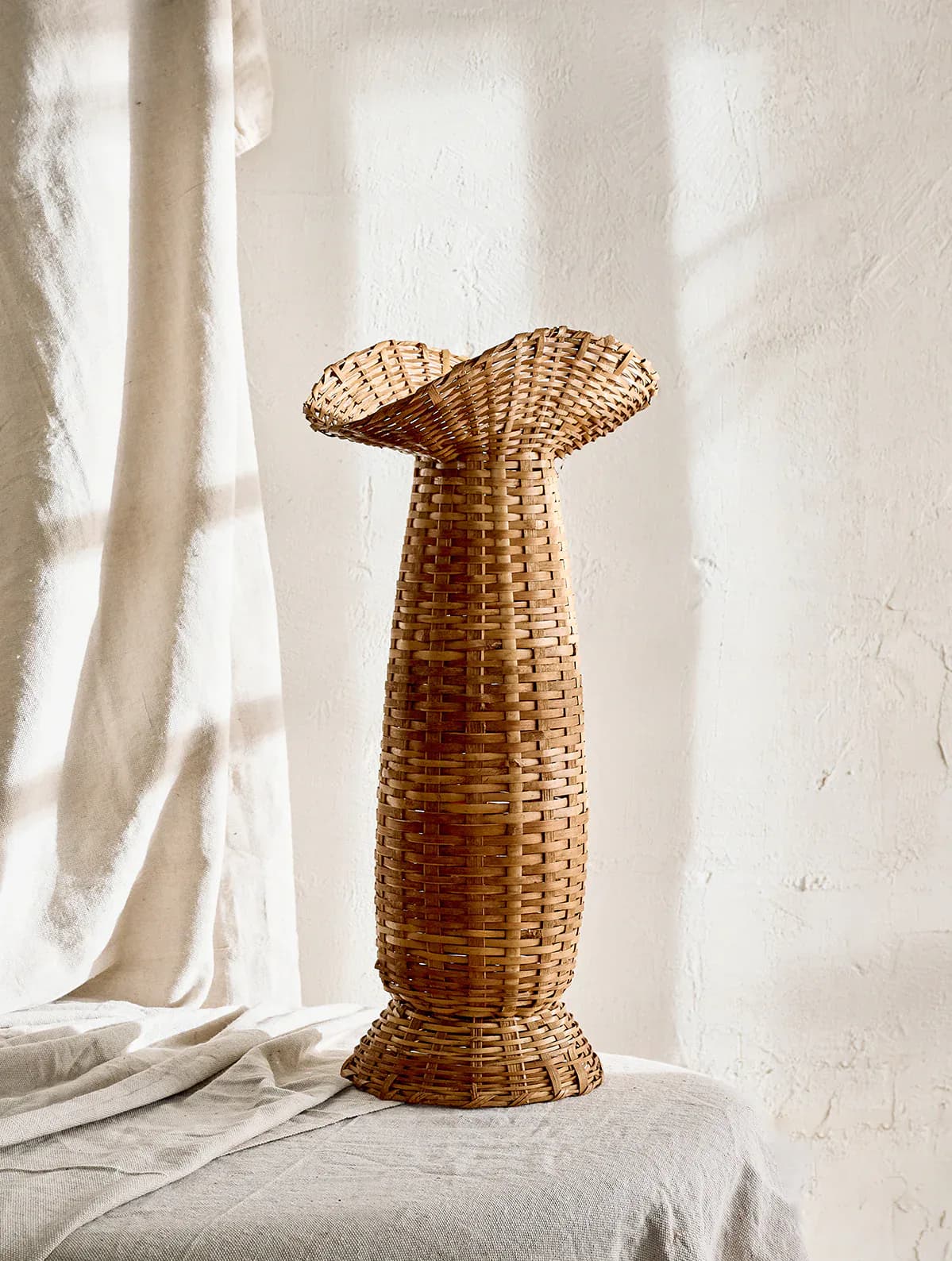 Woven wicker sculptural vessel with flared top displayed on linen fabric against white wall by SSS Atelier.