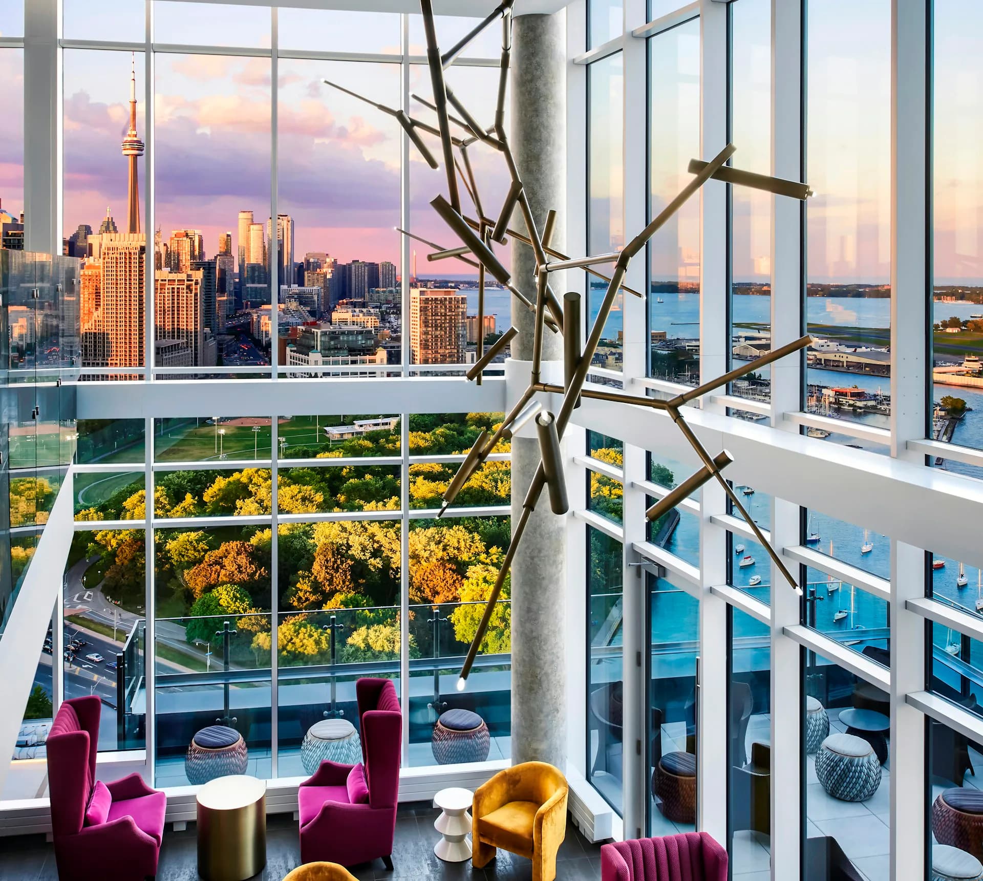 Modern high-rise lounge with floor-to-ceiling windows overlooking Manhattan skyline at sunset, featuring magenta and gold seating, sculptural branch installation, and panoramic water views by SBJ Group.