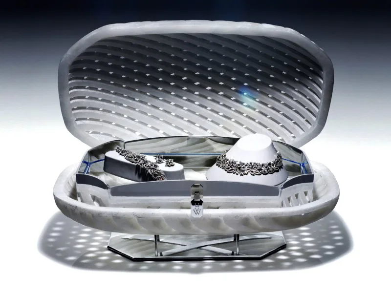 Futuristic white pod-shaped furniture piece with open lid revealing dark interior storage compartment and sculptural objects, rendered with studio lighting by Stephen Burks Man Made.