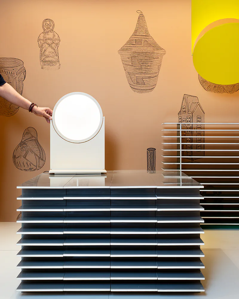Styled tabletop display featuring stacked navy and white books with a circular mirror, woven baskets, and wall drawings in warm earth tones by Stephen Burks Man Made.