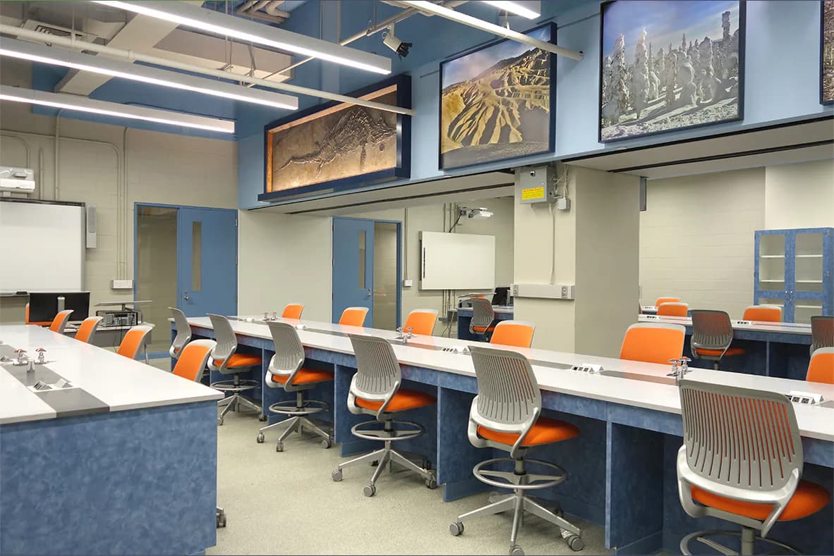 Modern computer lab with orange task chairs, blue and white cabinetry, white desks, and agricultural landscape artwork by Stephen Ely Architect.