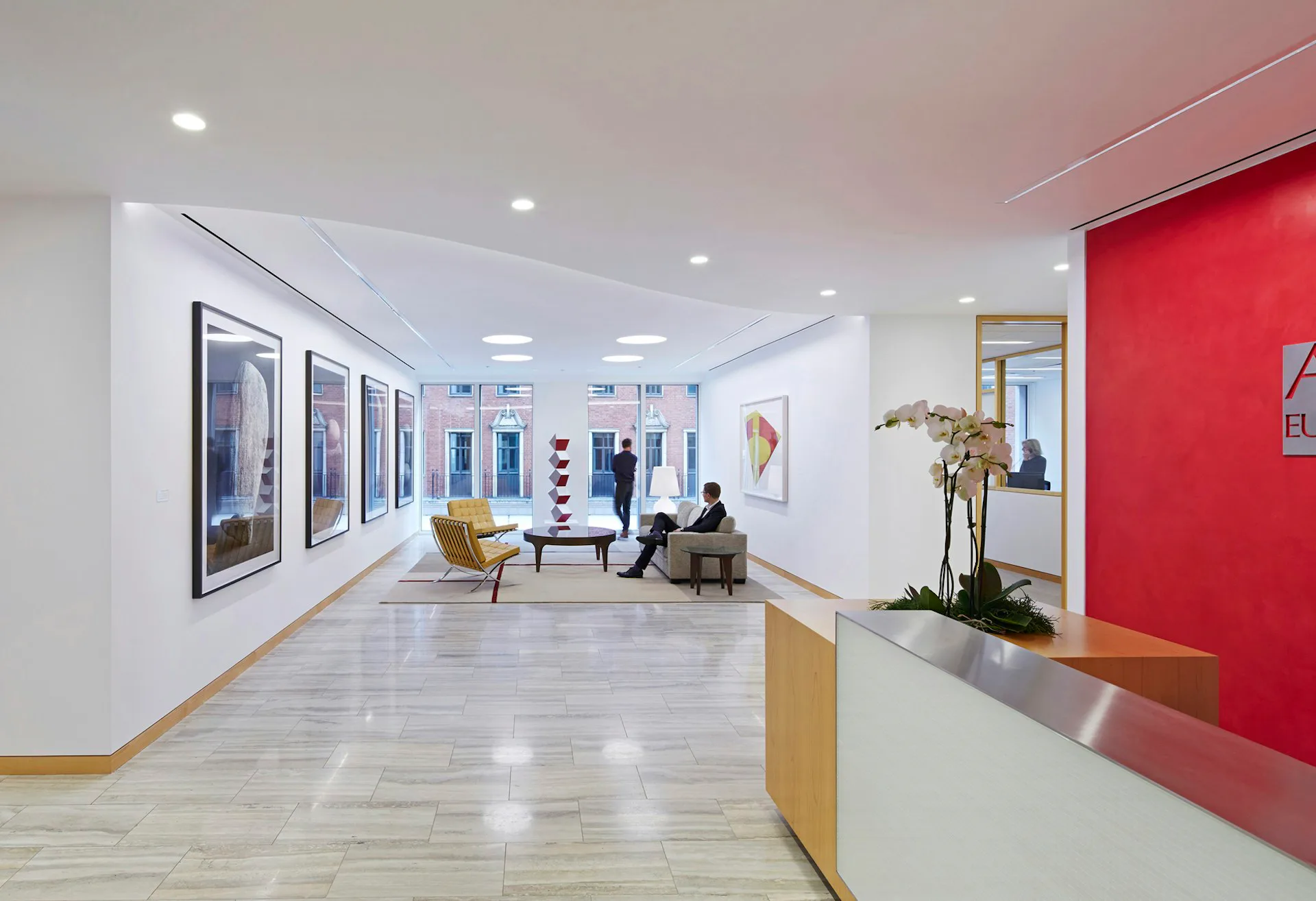 Modern medical office reception area with polished concrete floors, bright white walls, a bold red accent wall, wood reception desk, and stylized figures in the waiting area by Stephen Yablon Architecture.