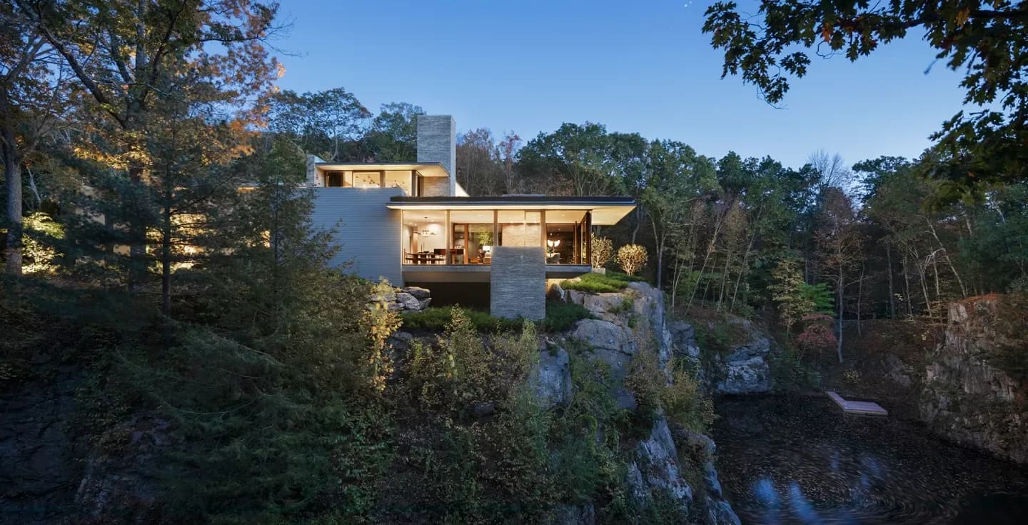 Modern residential structure with warm wood and glass facades perched on a rocky cliff overlooking a forested ravine, photographed at dusk by Steven Harris Architects.