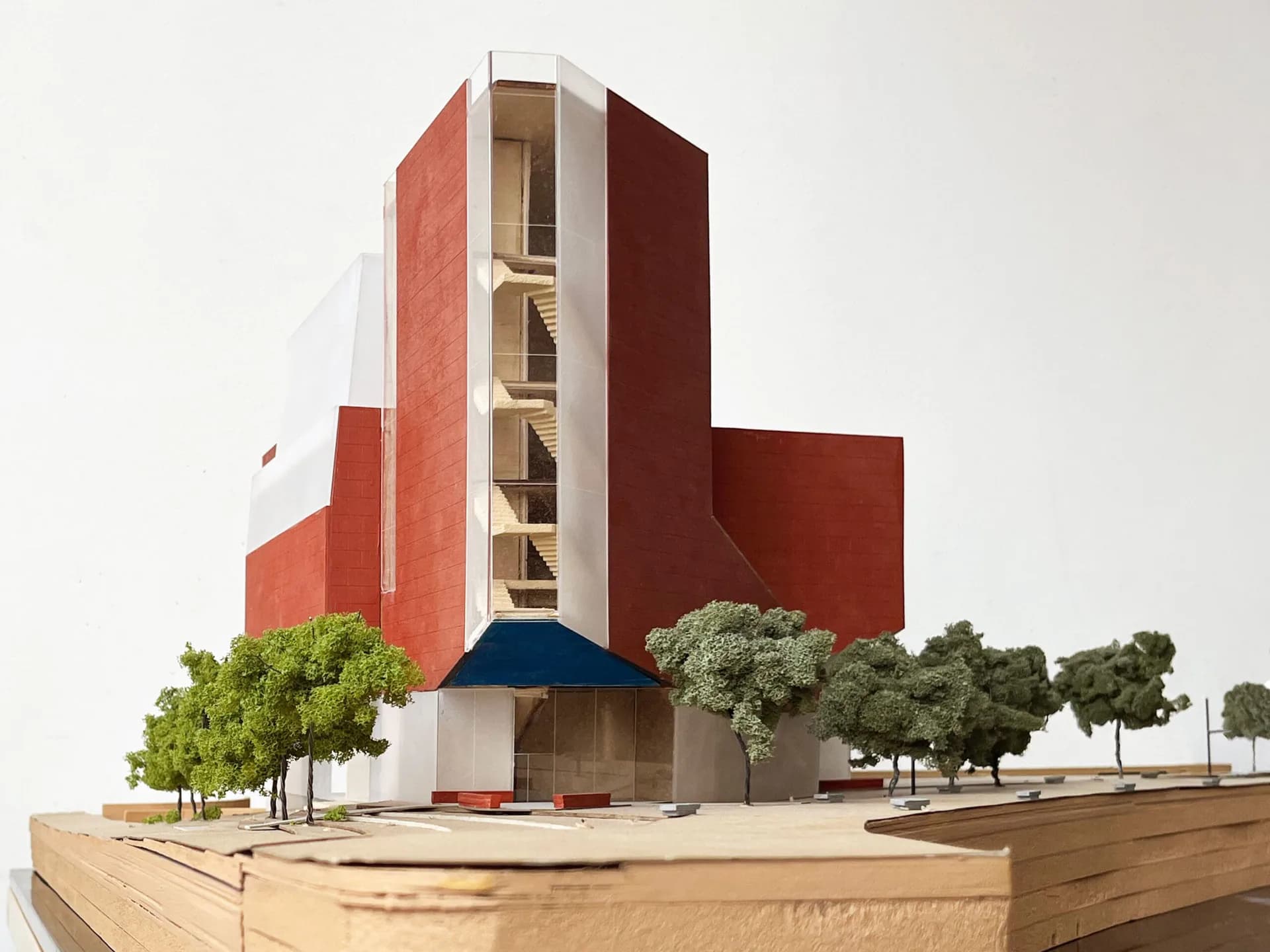 Architectural model of a red brick tower with white vertical core and deep blue canopy base, surrounded by landscaping trees on sandy terrain, by Steven Holl Architects.