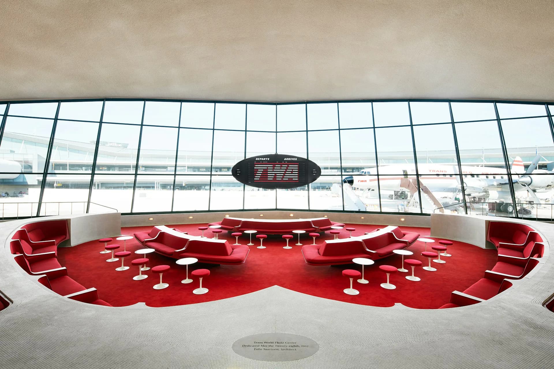 TWA Hotel's bright red seating installation beneath expansive gridded windows overlooking an airport runway, designed by Stonehill Taylor.