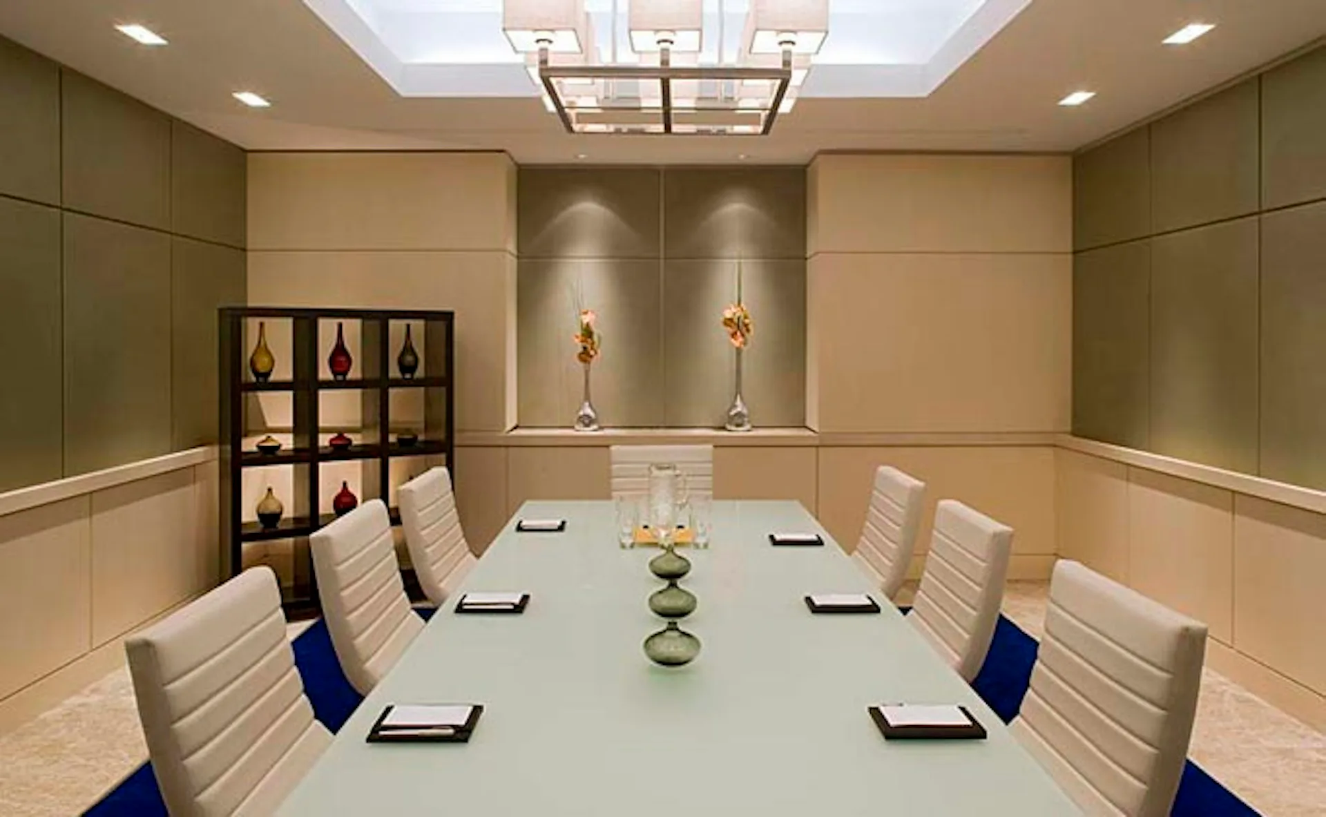 Modern conference room with light wood paneling, glass table, cream upholstered chairs, and built-in display shelving by Stonehill Taylor.