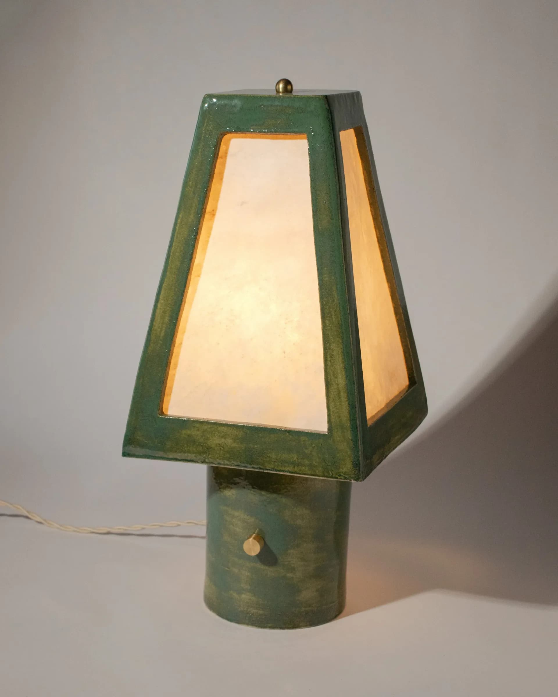 Green patinated metal table lamp with trapezoid shade and cream-colored panels, small brass sphere finial, lit from within by Streicher Goods.