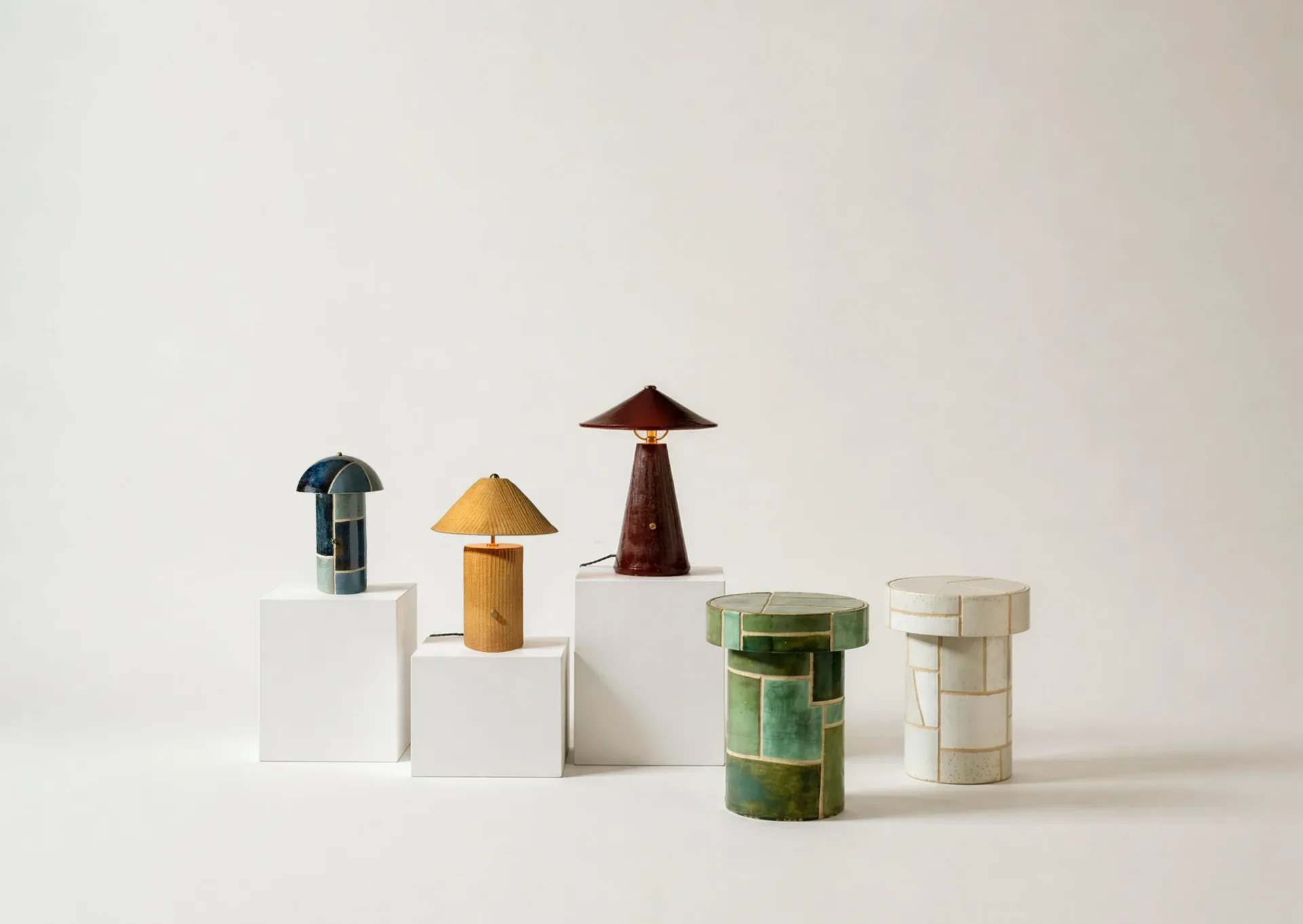 Stylized product arrangement featuring cylindrical vessels and geometric forms in teal, ochre, burgundy, and green tones on white platforms by Streicher Goods.