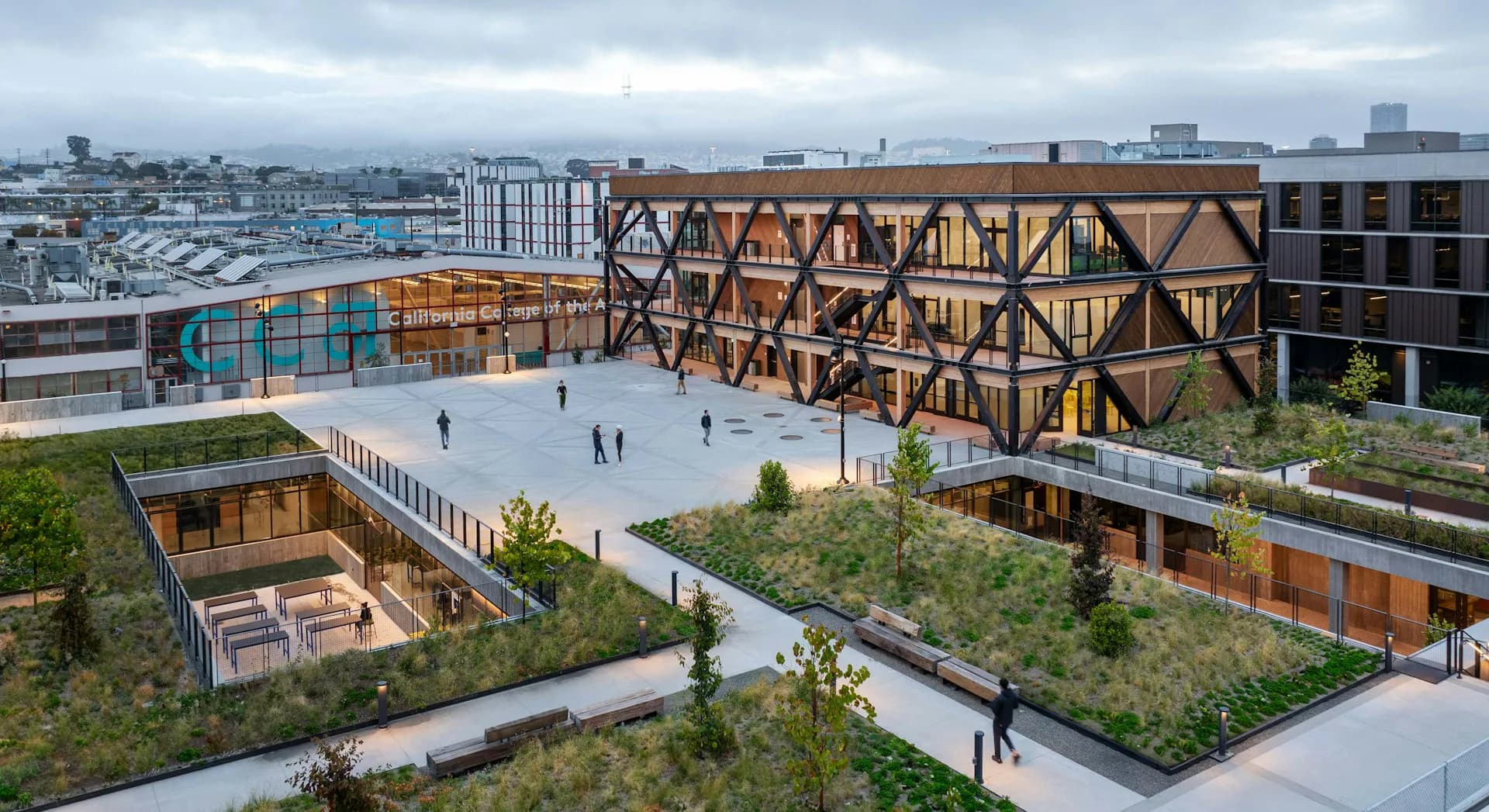 Aerial view of a mixed-use courtyard with timber-framed architecture, ground-level plaza with people, landscaped green spaces, and city skyline beyond by Studio Gang.