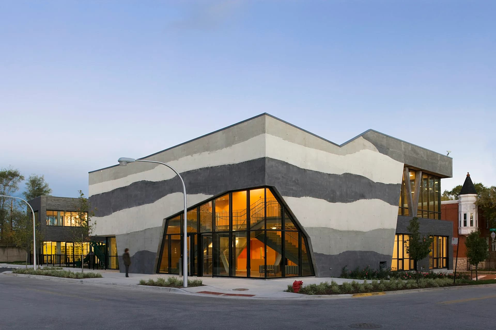 Contemporary building with sculptural wavy concrete facade and large orange-tinted glass windows at dusk, designed by Studio Gang.