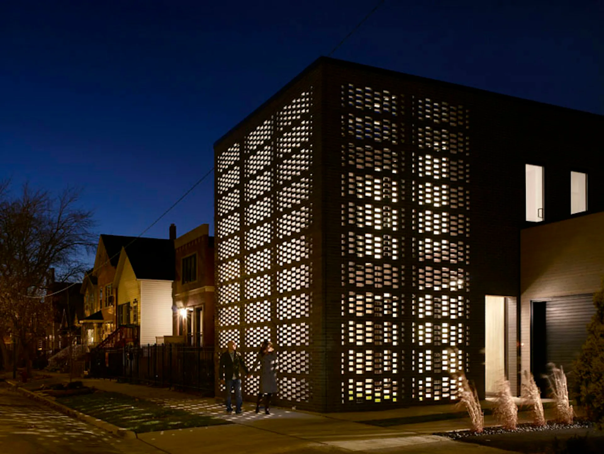 Modern brick building with illuminated geometric lattice screen facade at dusk, featuring strip windows and minimalist design by Studio Gang.