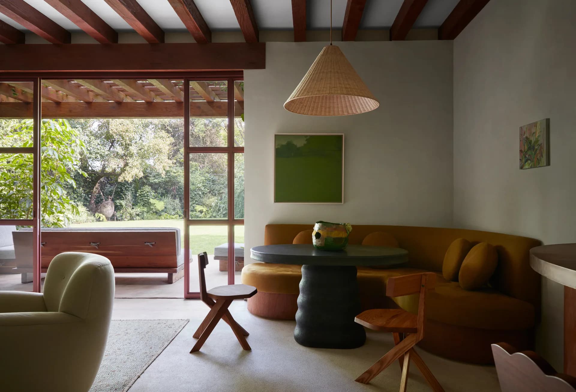 A mid-century modern living room with exposed wooden beams, cognac leather sofa, woven pendant light, and large windows overlooking greenery by Studio Valle de Valle.