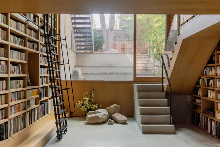 Warm ochre library loft with floor-to-ceiling bookshelves, floating wooden mezzanine, metal ladder, and large windows overlooking urban landscape by ERGO Architects.