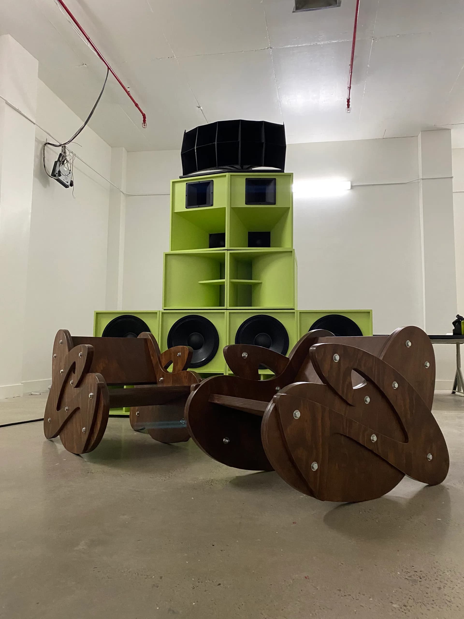 Contemporary art installation featuring lime-green cubic shelving tower with black circular elements and brown sculptural wheel-like forms on polished concrete floor by Studio Guapo.