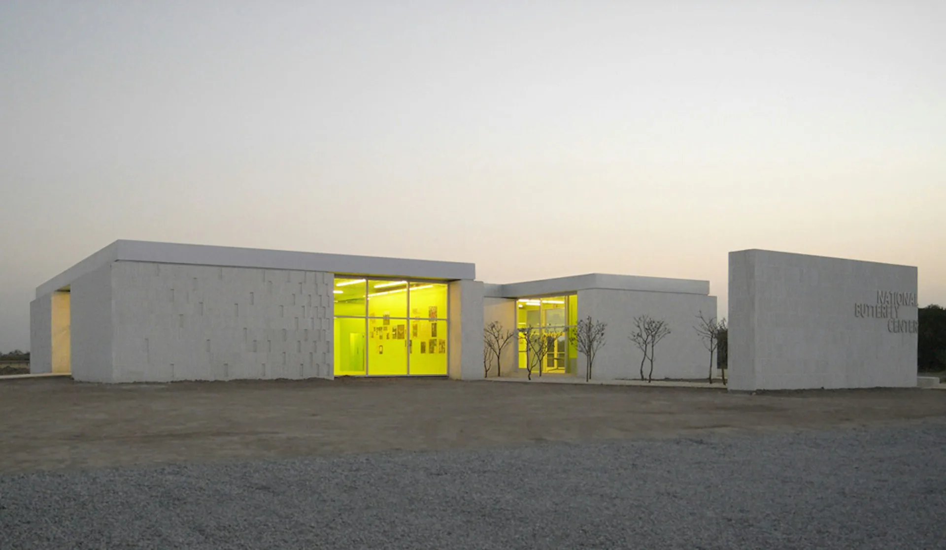 Modular concrete pavilions with illuminated yellow-green interior spaces and bare trees against twilight sky by Studio Joseph.