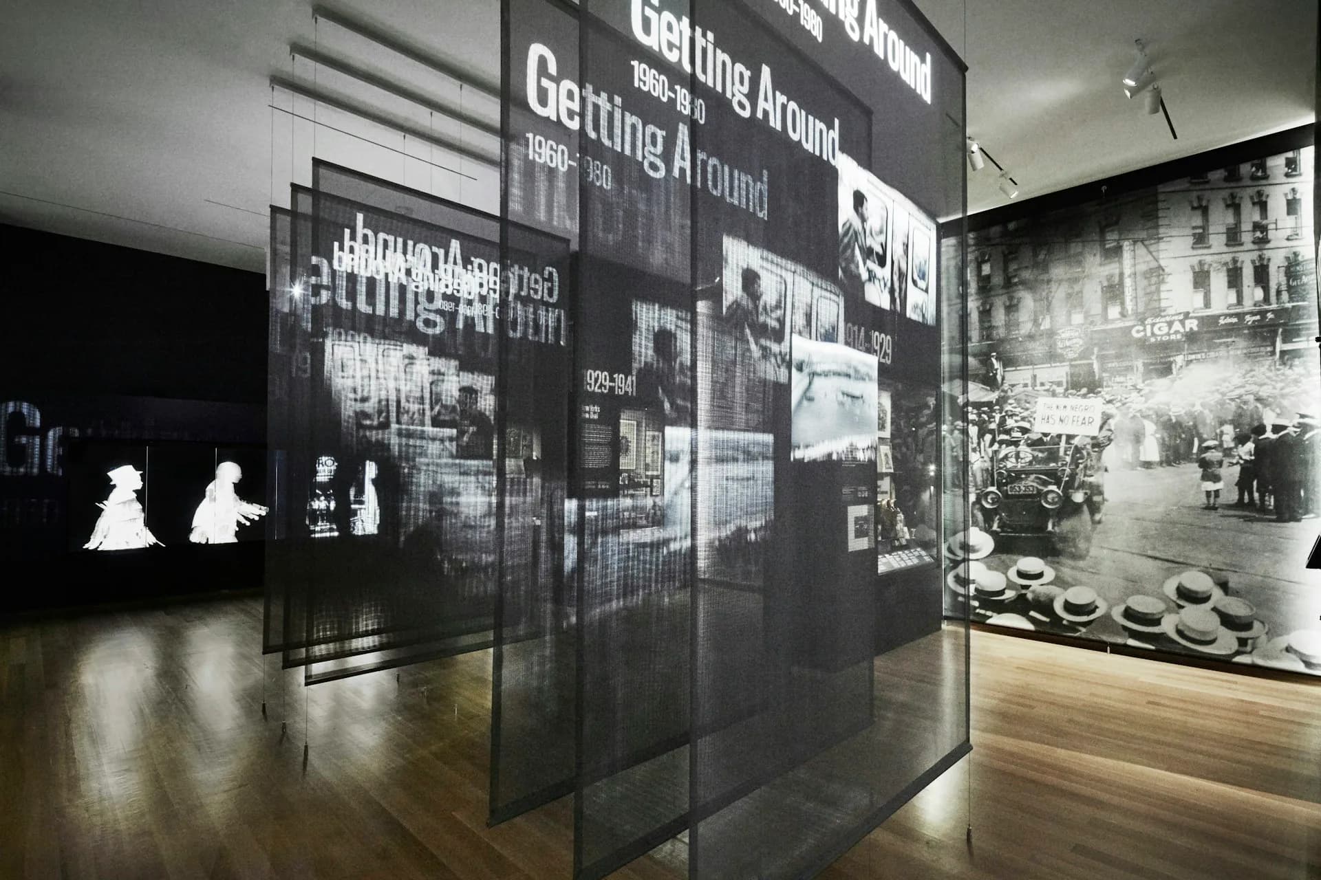 Dark exhibition interior with translucent display panels featuring historical photographs and text, wooden flooring, and ambient lighting creating a contemplative museum space by Studio Joseph.