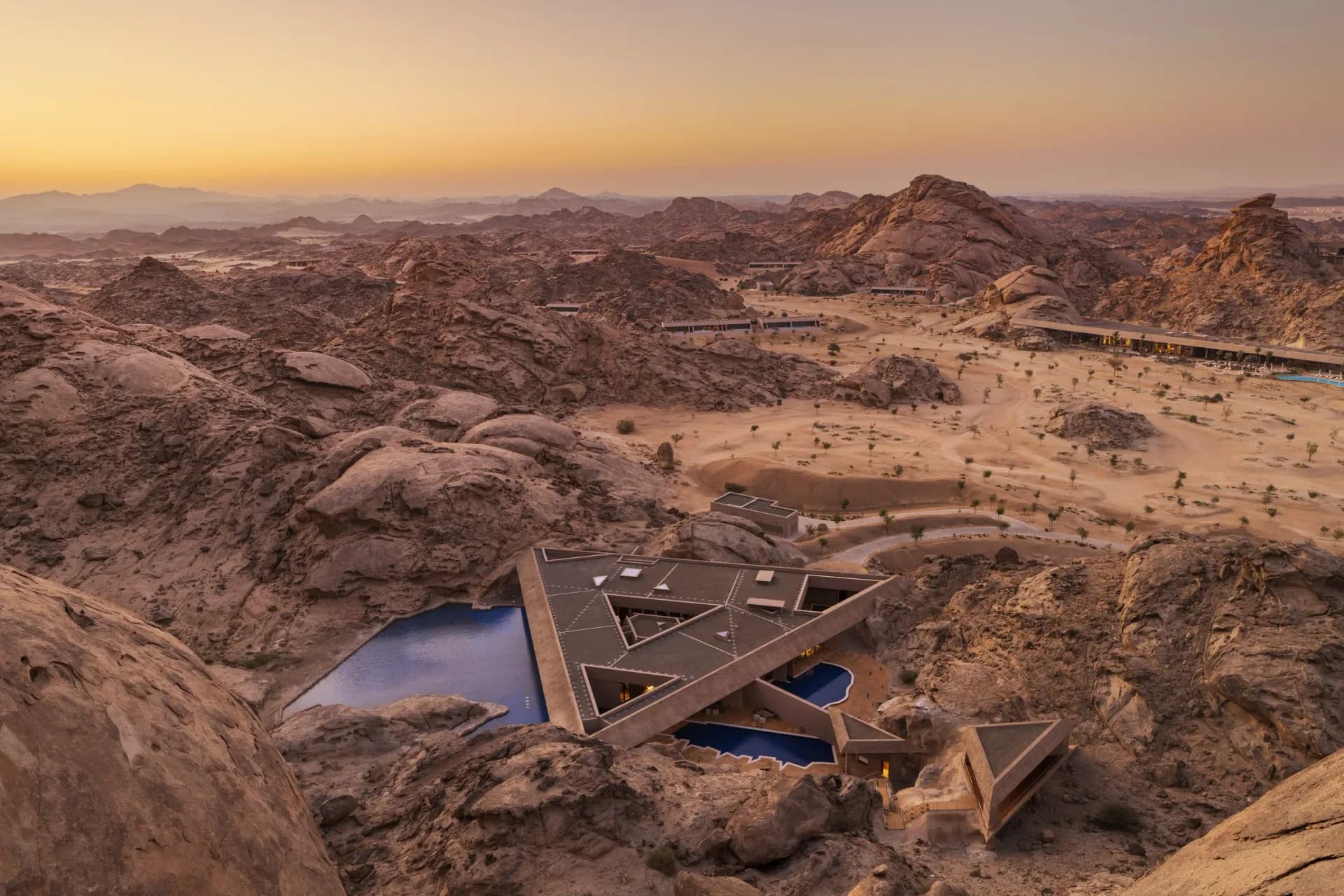 Aerial view of a contemporary geometric structure with blue pools nestled in desert landscape at sunset by Studio Paolo Ferrari.