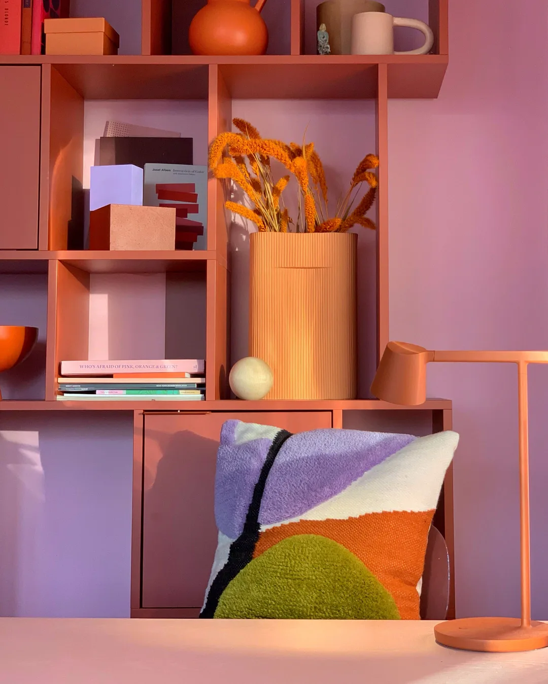 Coral-pink modular shelving unit with geometric storage displaying dried flowers, books, and decorative objects against a lavender wall by Studio Proba.