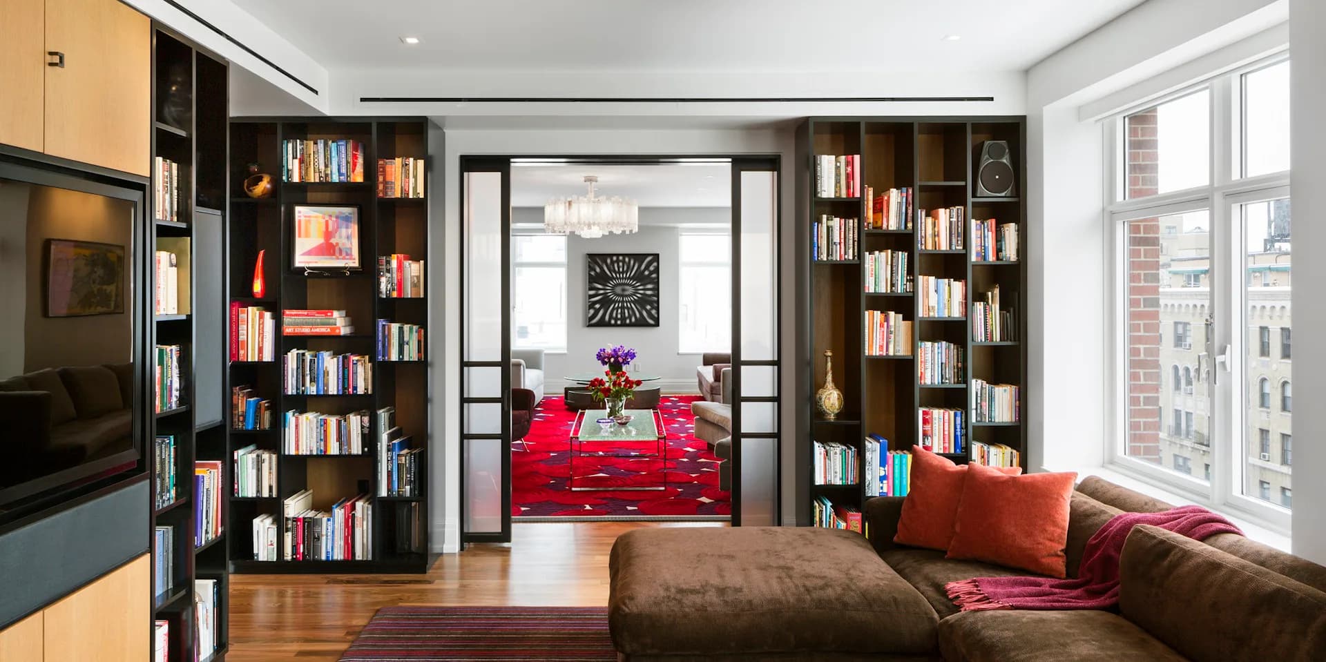 Modern library lounge with floor-to-ceiling wooden bookshelves, red velvet seating, brown leather sofa, and large windows, designed by Studio ST Architects.