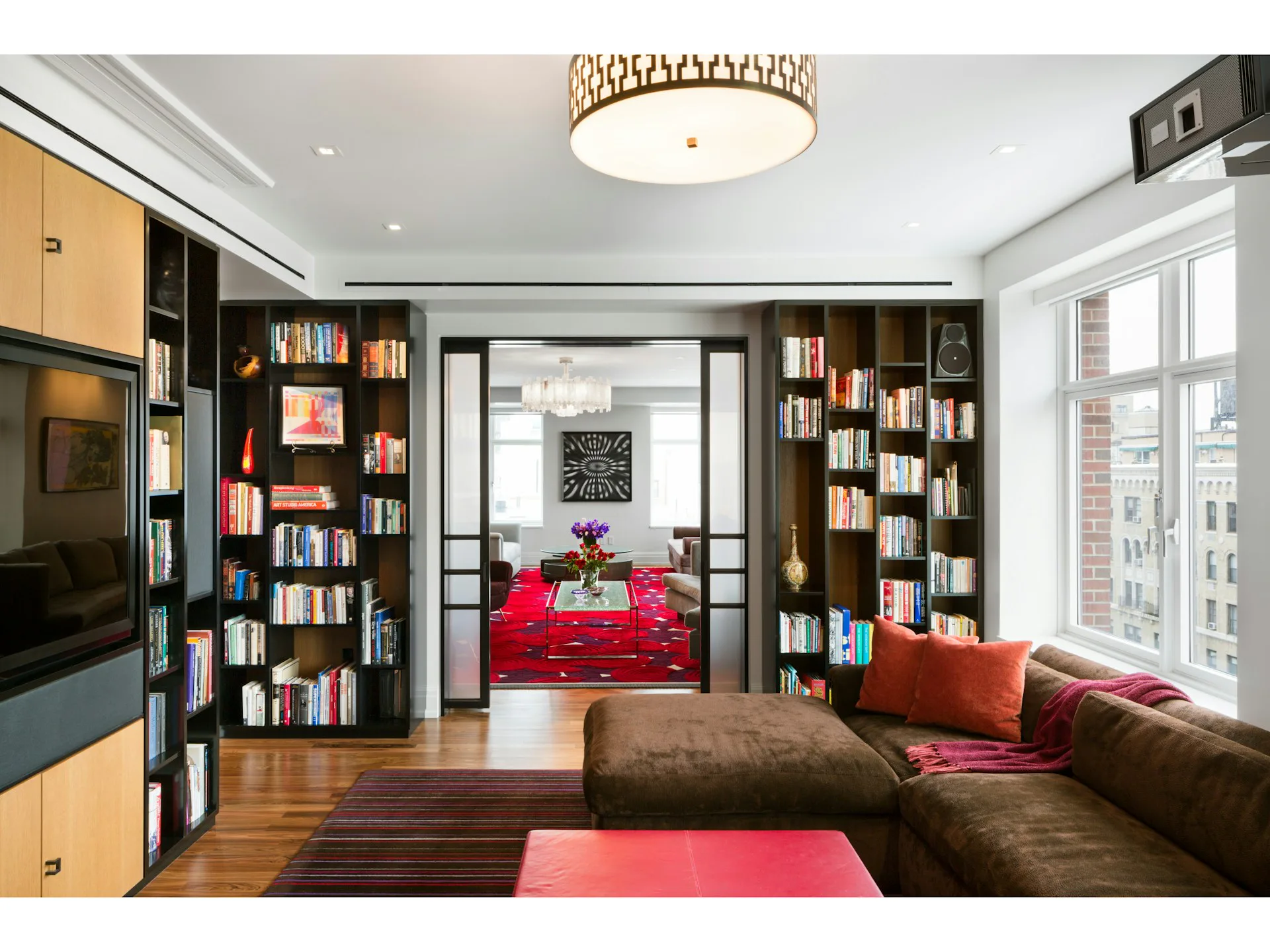 Modern library lounge with built-in bookshelves, warm wood flooring, vibrant pink seating, and glass doors opening to a bedroom beyond, by Studio ST Architects.