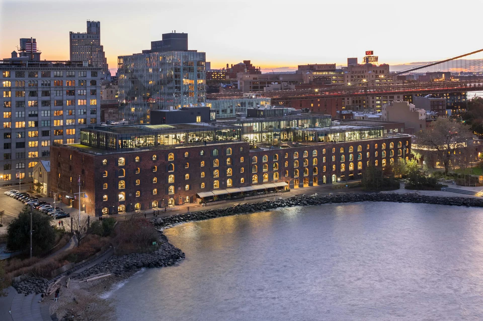 Waterfront residential building with illuminated facade at dusk, featuring repetitive window pattern overlooking water and cityscape by STUDIO V.