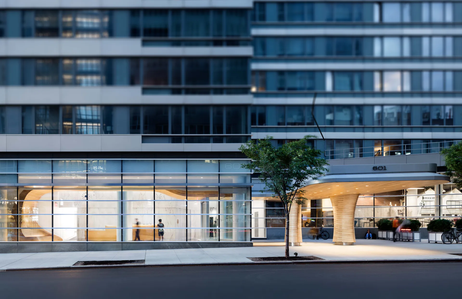 Modern ground-floor retail and office space with floor-to-ceiling glazing, warm interior lighting, minimalist furnishings, and a potted tree by STUDIO V.