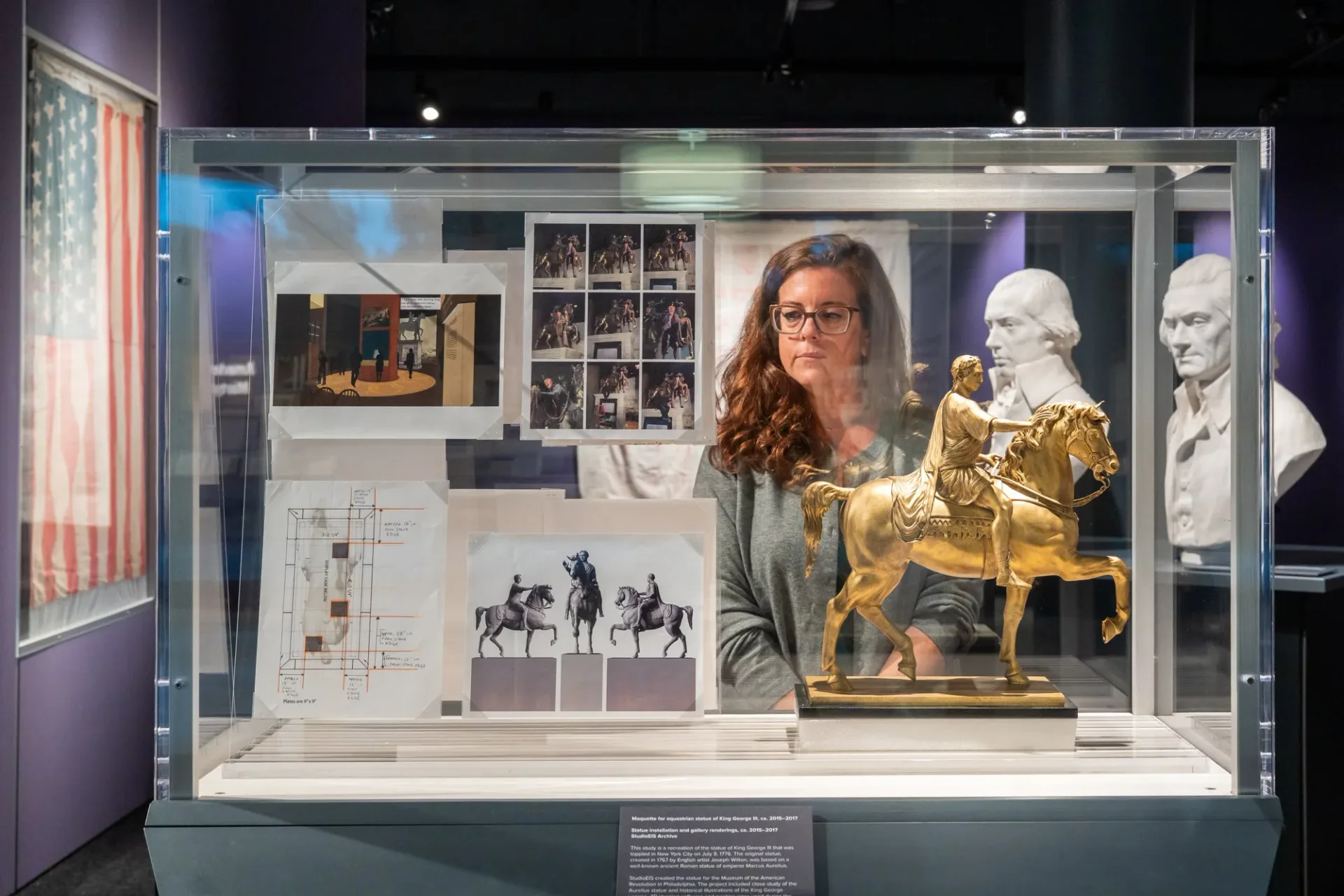 Museum exhibition display case featuring archival materials, photographs, and golden sculptural elements with explanatory panels, designed by StudioEIS.