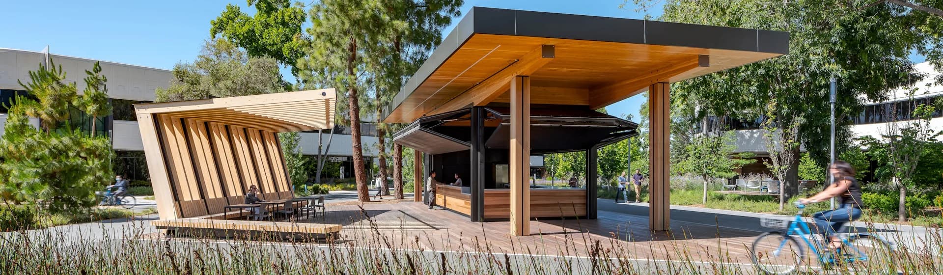 Modern wooden pavilion with metal roof and integrated seating area in a landscaped park setting, designed by STUDIOS Architecture.