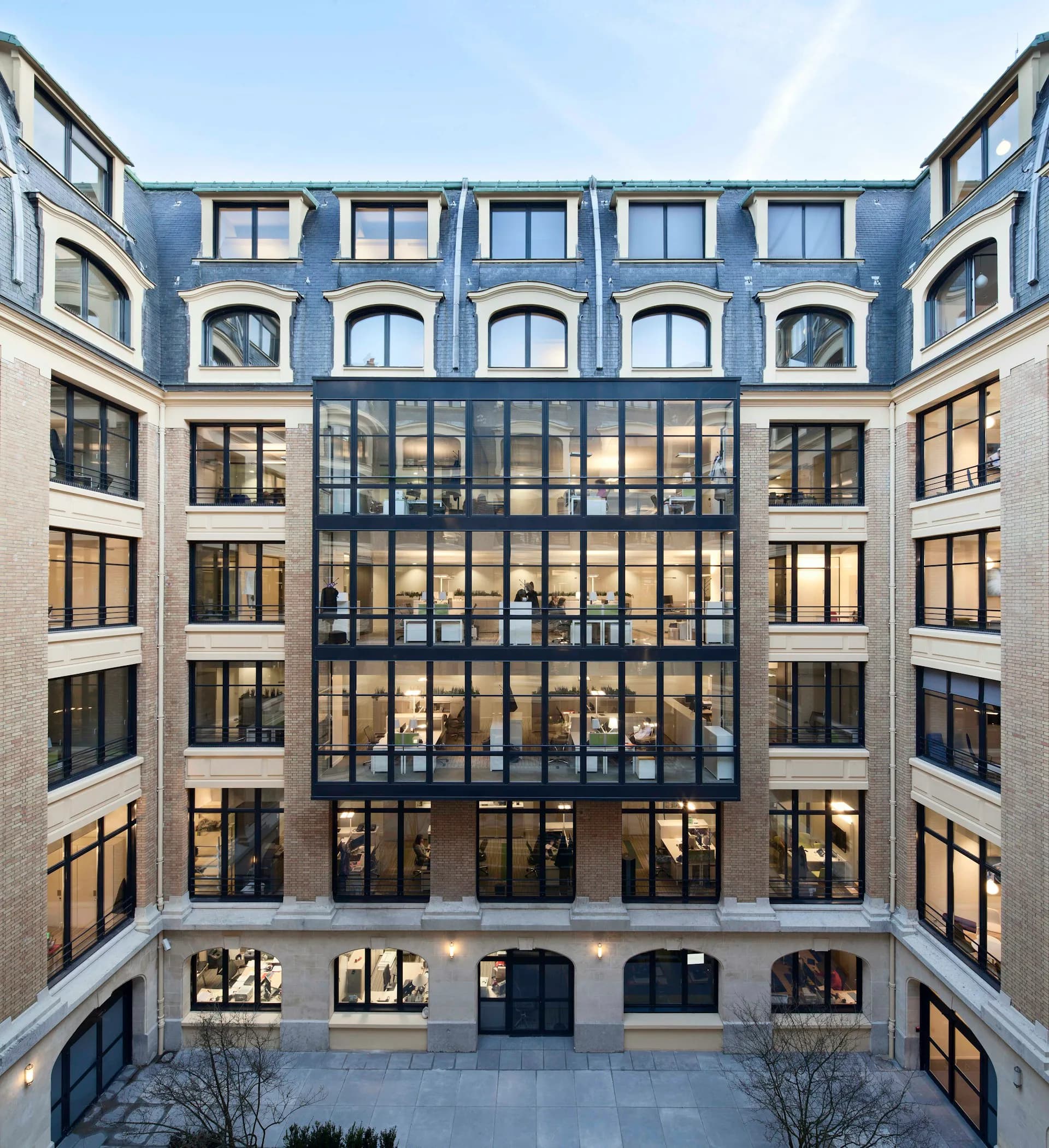 A modern Parisian courtyard facade featuring symmetrical cream and blue stone buildings with mansard roofs and a central glass and steel atrium structure, designed by STUDIOS Architecture.
