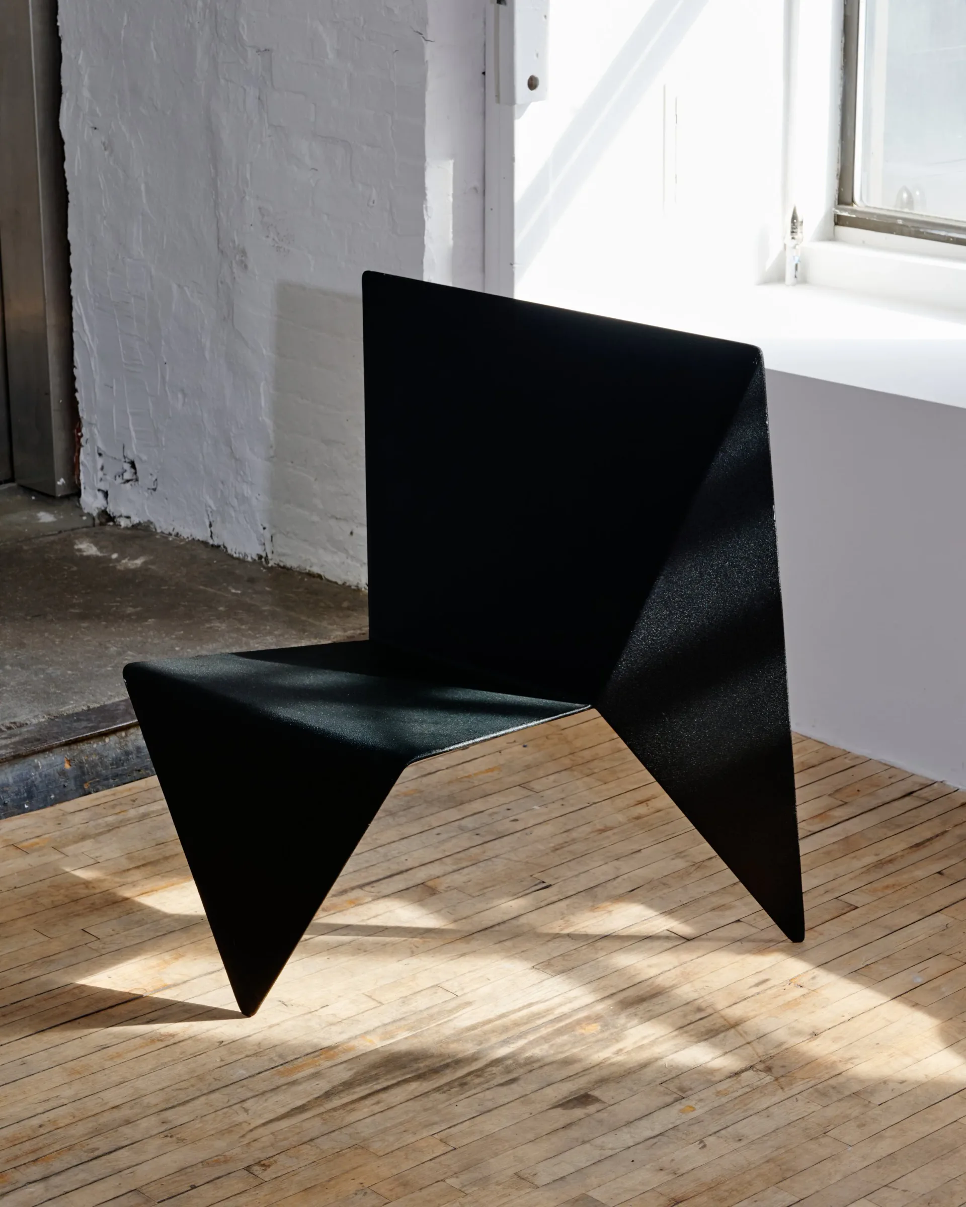 Black sculptural chair with angular geometric forms sits on light wood flooring in bright studio space with white walls, by Superhouse.