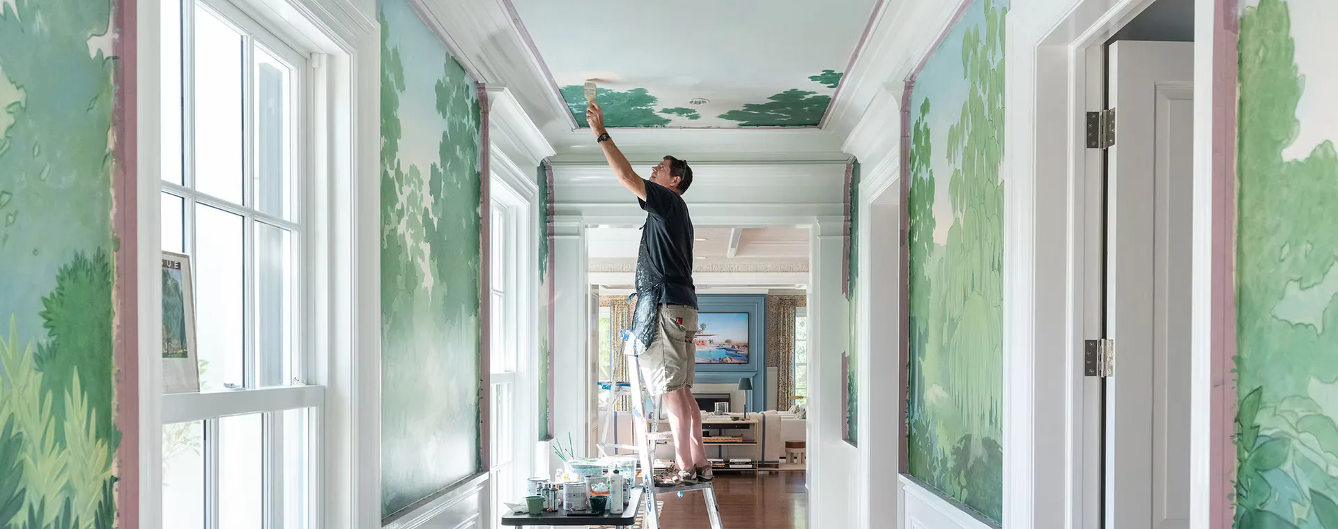 Hallway with green botanical wall murals, natural light from large windows, person on ladder painting ceiling, modern residential interior by SuperStrata.
