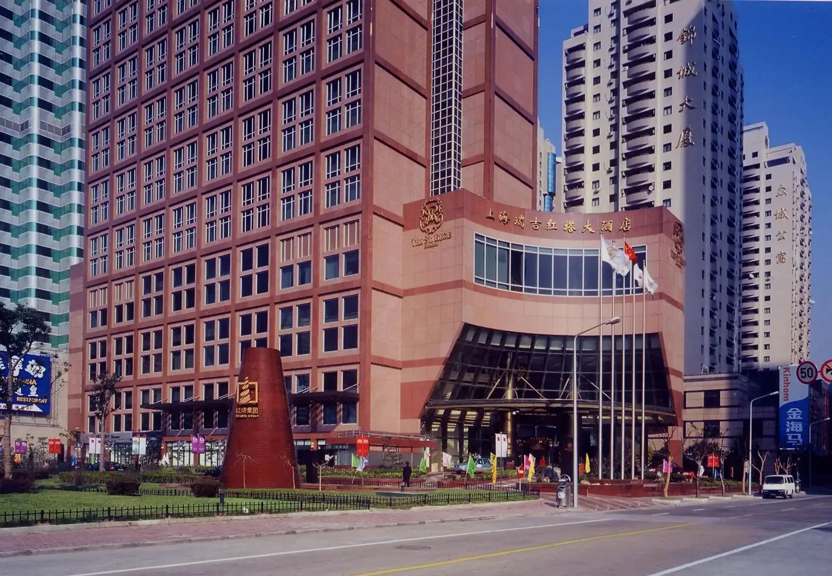 Red brick and glass modernist building with curved facade and plaza foreground, surrounded by tall residential towers under clear blue sky, by Sydness Architects.