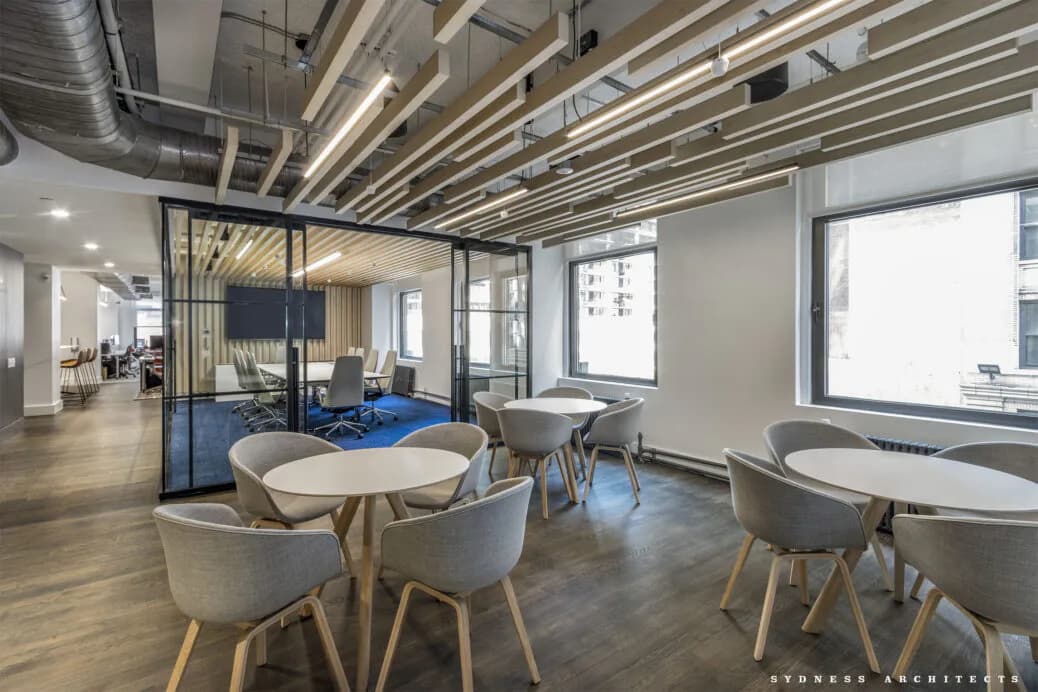 Modern office break room with round tables, gray chairs, wood flooring, and floor-to-ceiling windows by Sydness Architects.