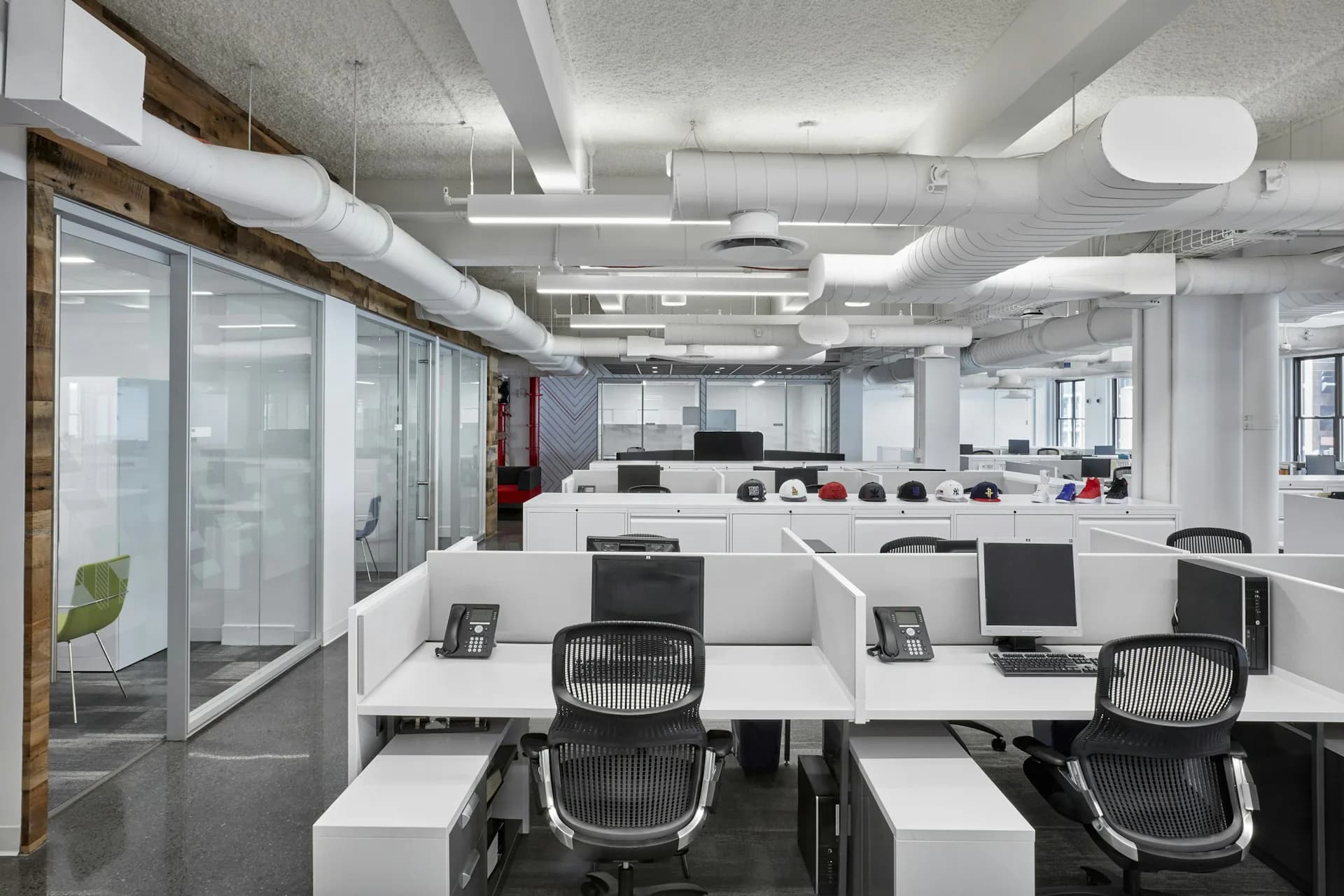 Modern open-plan office with white workstations, black ergonomic chairs, glass partitions, and industrial ceiling details by Ted Moudis Associates.