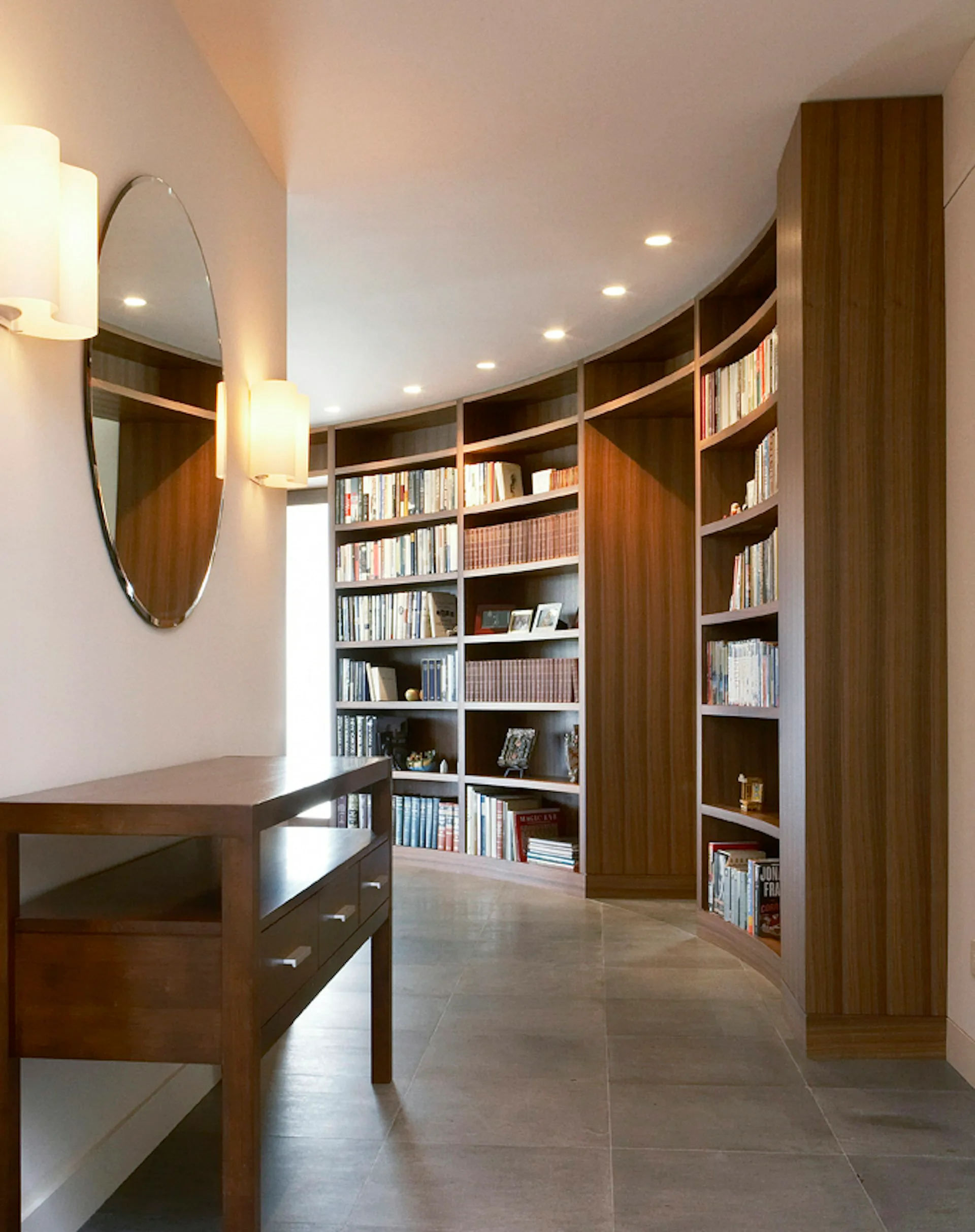 Modern library corridor with curved wooden shelving units, warm wood tones, recessed lighting, polished concrete floors, and a desk with mirror by Ted Porter Architecture.