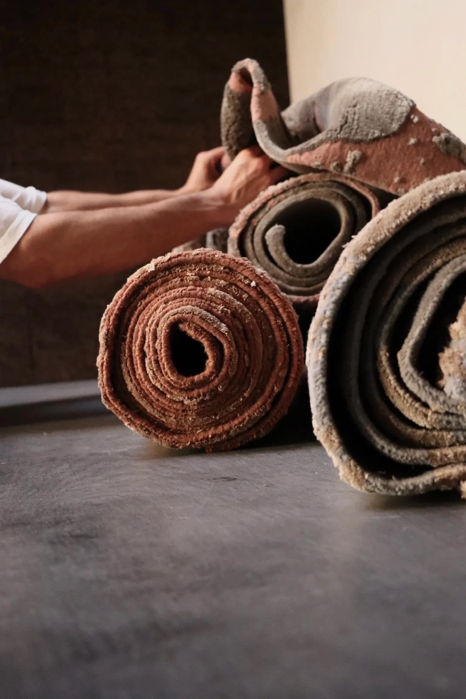 Rolled rugs in warm earth tones—rust, cream, and charcoal—displayed on concrete flooring with a hand touching the textured surface by Temple Rugs.