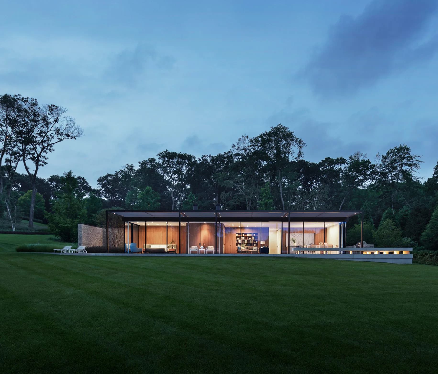 Modern minimalist residential building with extensive glazing sits within expansive lawn surrounded by mature trees at dusk, illuminated interiors visible by TenBerke.