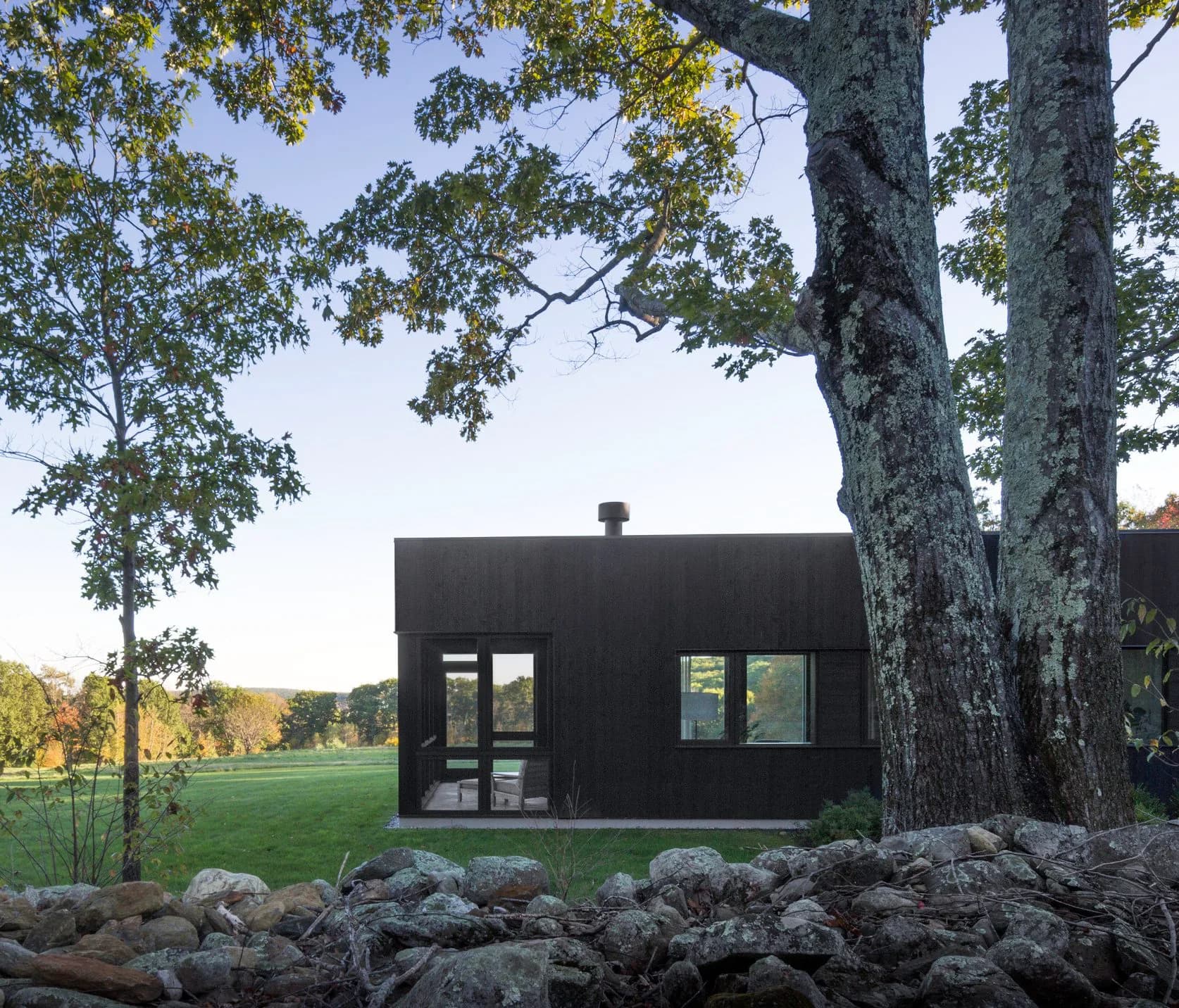 Modern black residential pavilion with large windows nestled beneath mature trees in a green landscape setting by TenBerke.