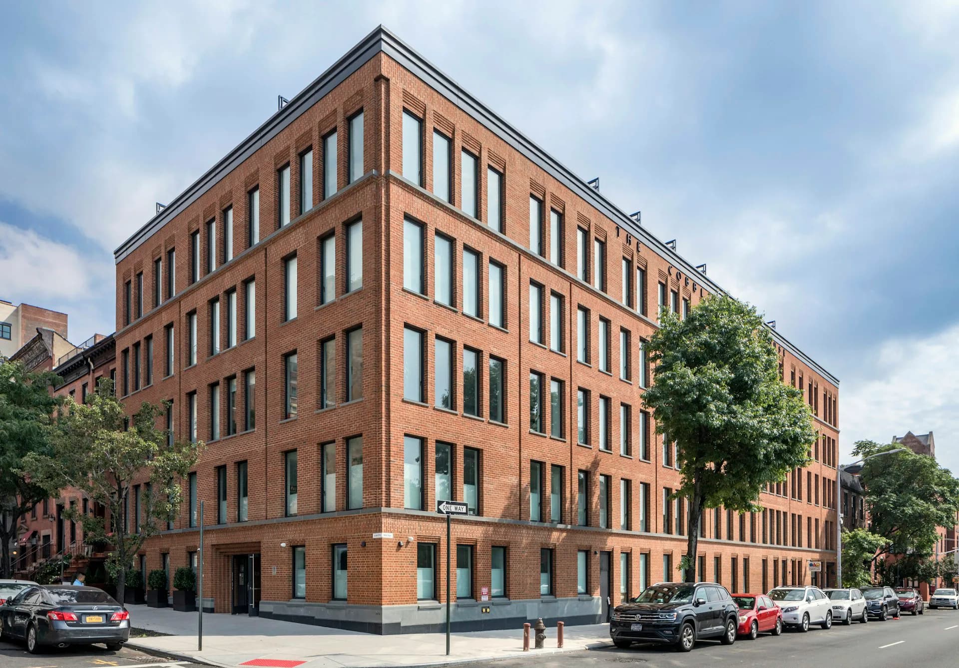 Corner view of a restored red-brick residential building with regular window patterns, tree-lined street frontage, and ground-floor retail spaces by The Brooklyn Studio.