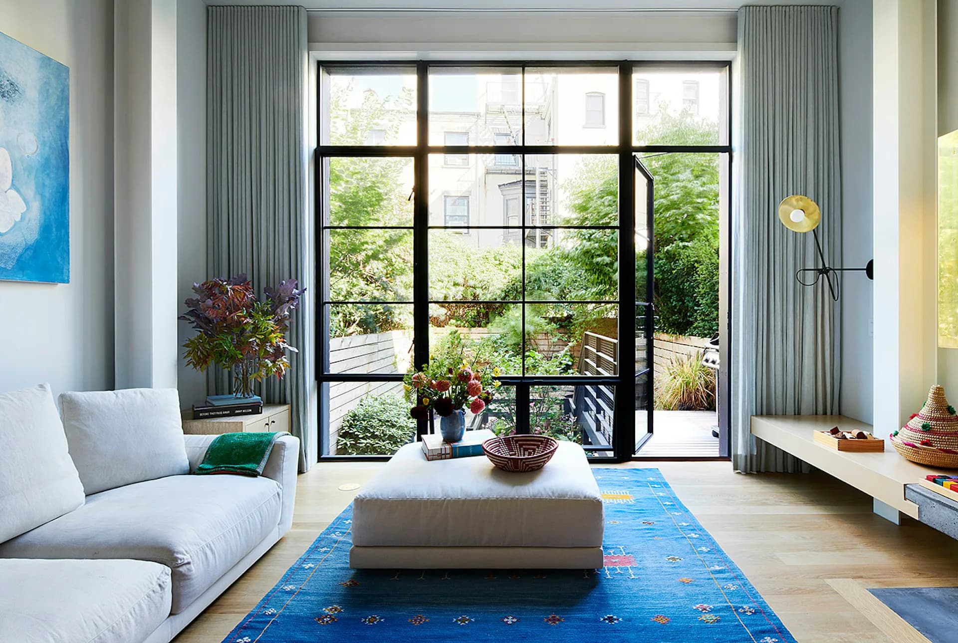 Bright contemporary living room with floor-to-ceiling black-framed glass doors, gray upholstered seating, blue geometric rug, and lush green views by The Brooklyn Studio.