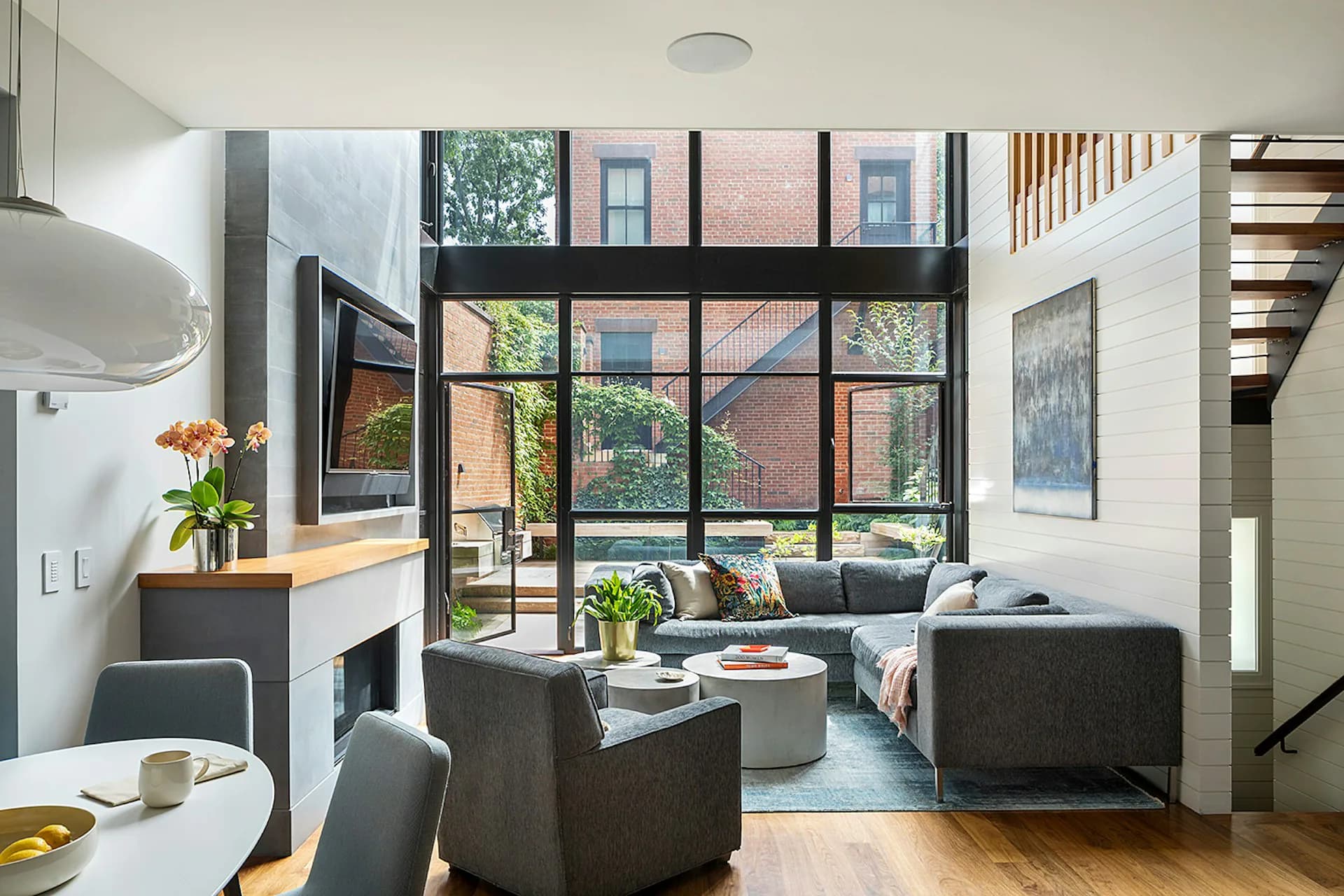 Bright loft living room with charcoal sectional, large black-framed windows overlooking brick buildings, wood flooring, and minimalist furnishings by The Brooklyn Studio.