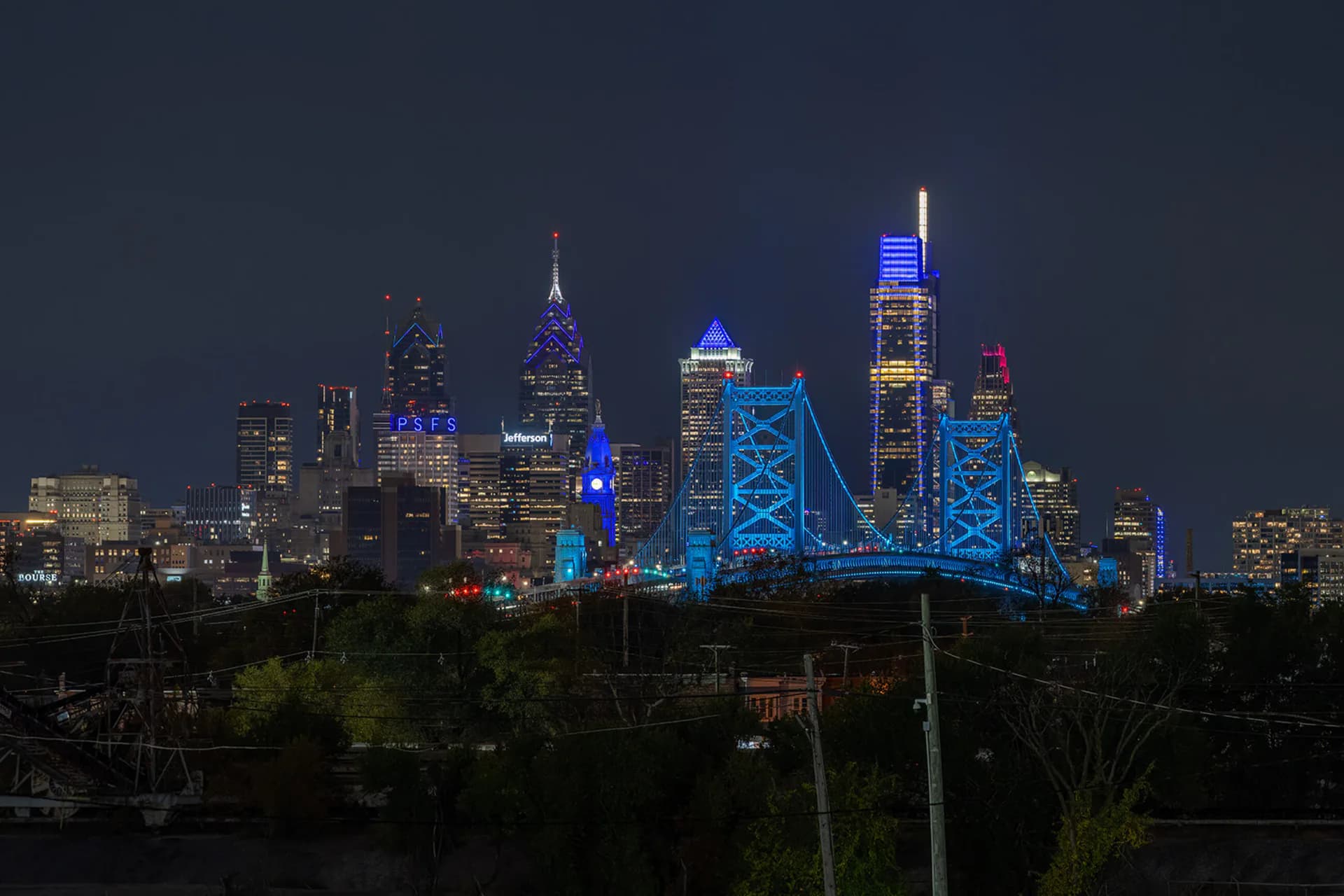 Manhattan skyline at night with architectural lighting design illuminating iconic skyscrapers and the Queensboro Bridge in vibrant blue tones by The Lighting Practice.