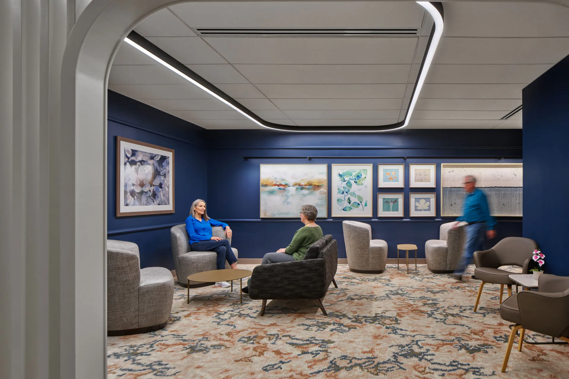 Modern office lounge with deep blue walls, patterned carpet, and contemporary furniture arrangement featuring multiple seating areas and artwork by The Lighting Practice.