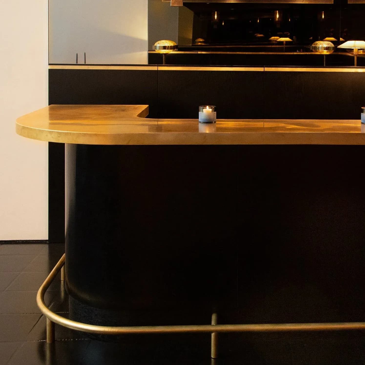 Curved bar counter with warm wood top and brass rail detail, black facade, ambient lighting above, minimalist design by The New Motor.