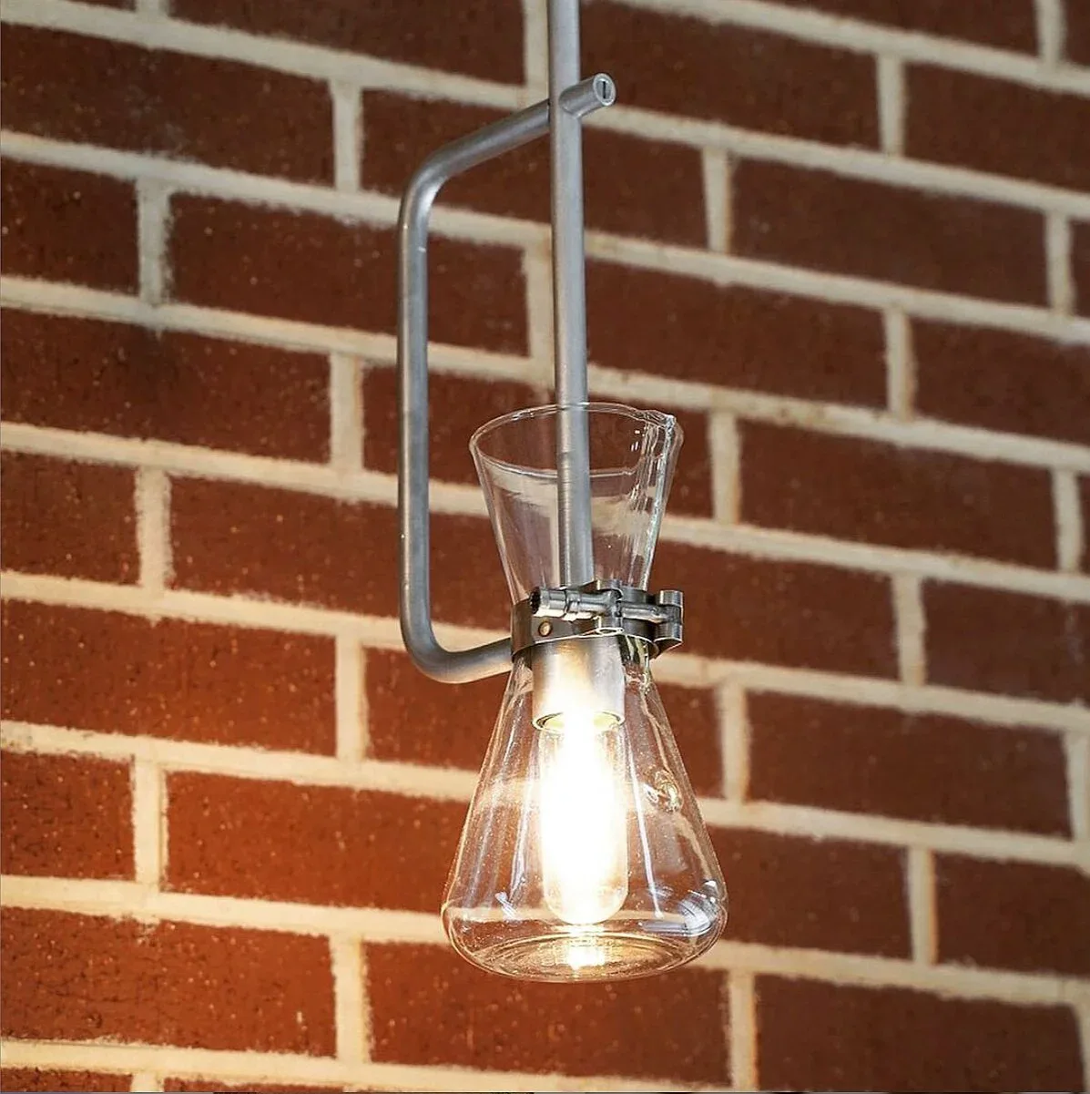 Minimalist pendant light fixture with clear glass shade and curved metal arm mounted against brick wall by The New Motor.