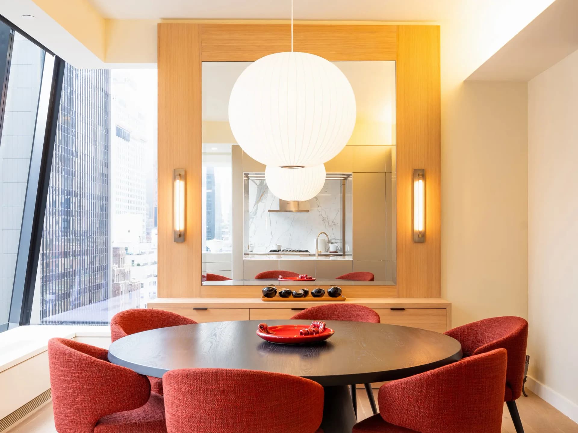 Modern dining room with curved black table, coral red chairs, and large white pendant light, warm wood surround and wall sconces by The New Motor.