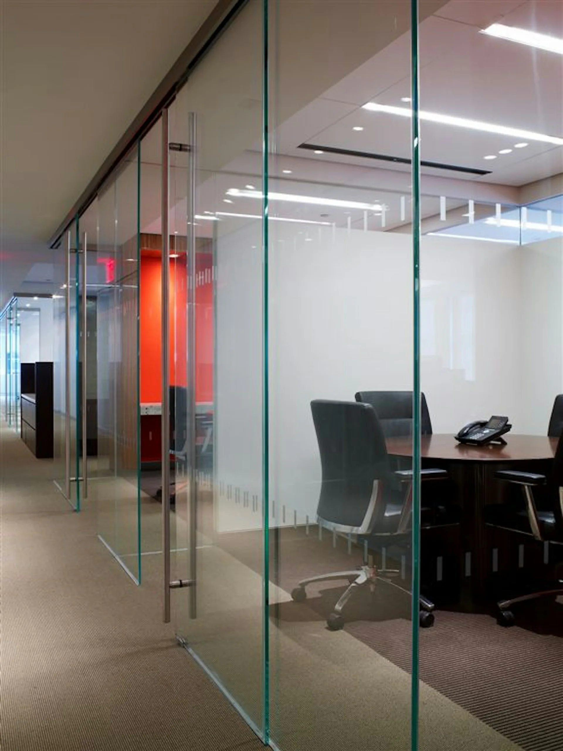 Modern glass-partitioned office space with frosted panels, black office chair, and red accent wall visible through transparent divisions by The Switzer Group.
