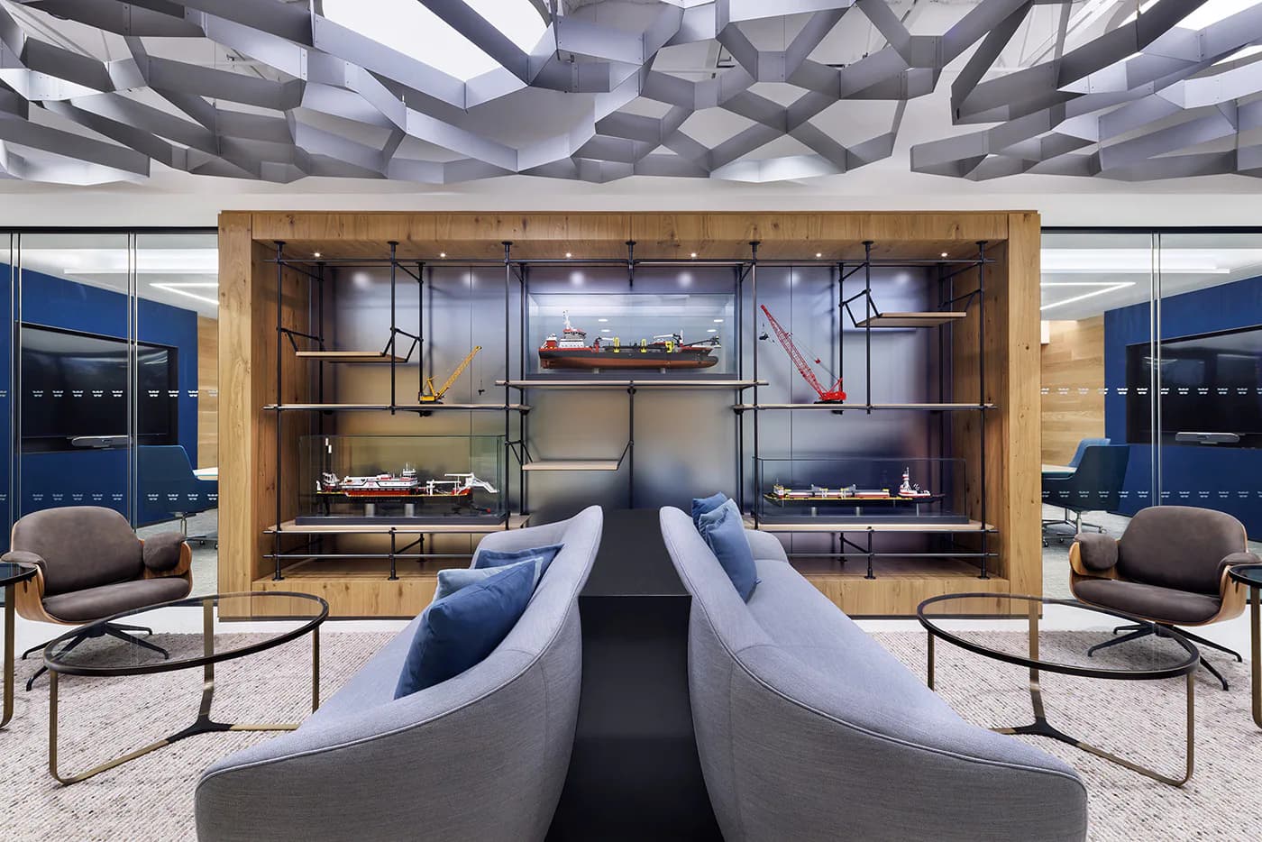 Modern office lounge with symmetrical curved seating, wooden wall display cases, geometric ceiling panels, and floor-to-ceiling windows overlooking a cityscape by The Switzer Group.