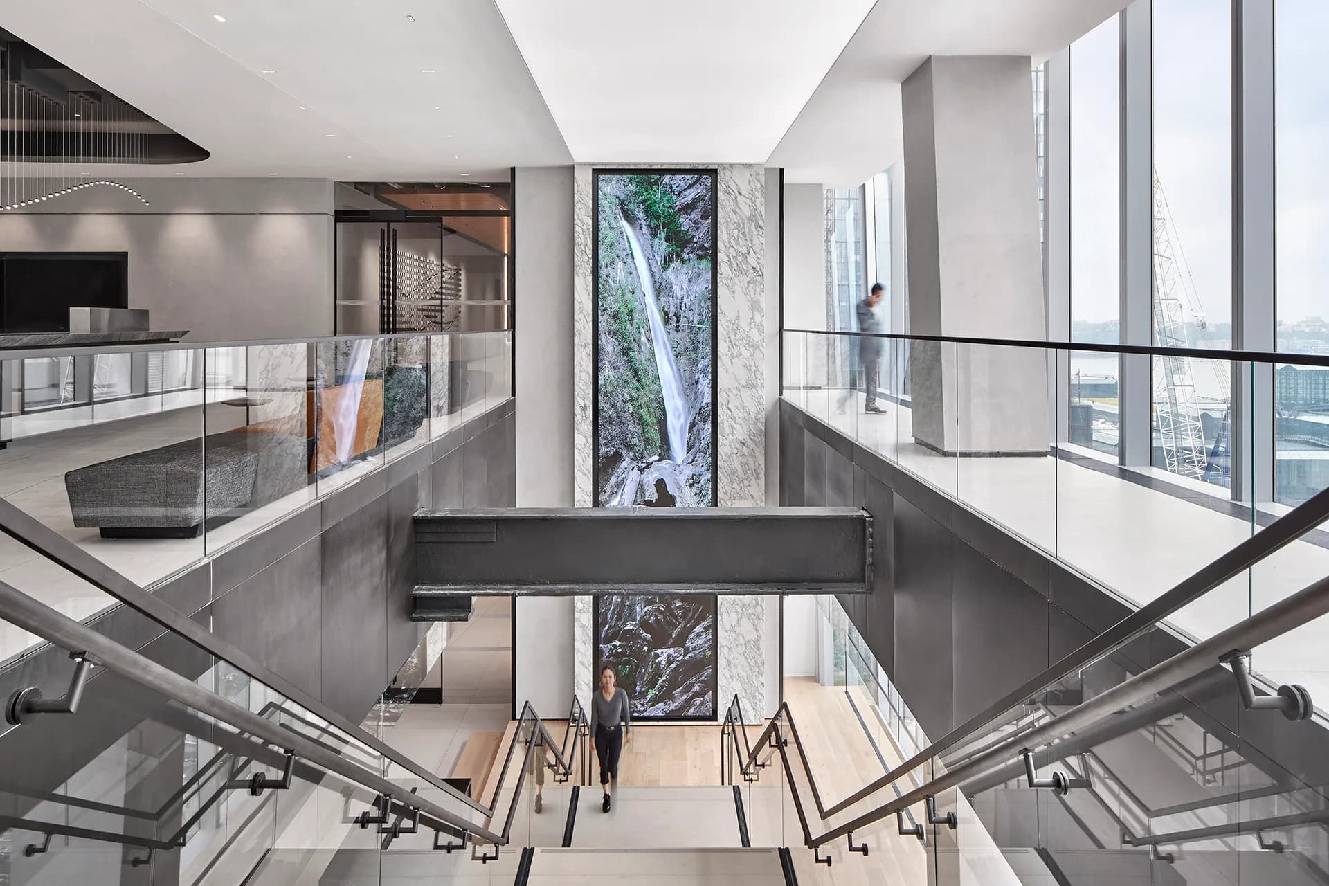 A modern multi-story atrium with glass railings, polished concrete floors, and floor-to-ceiling windows overlooking trees, featuring a minimalist aesthetic by The Switzer Group.