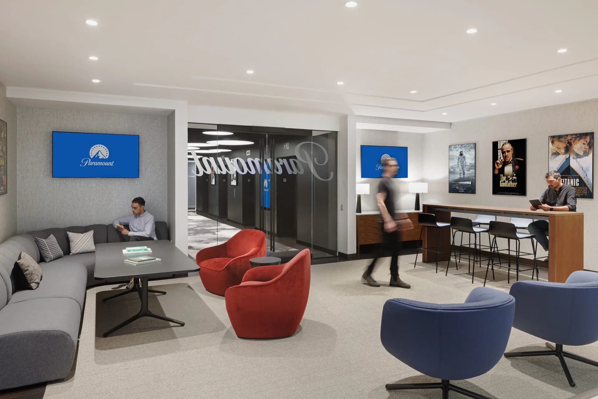 Modern commercial lounge interior with gray and blue seating, red accent chairs, wood cabinetry, digital displays, and polished concrete flooring by The Switzer Group.